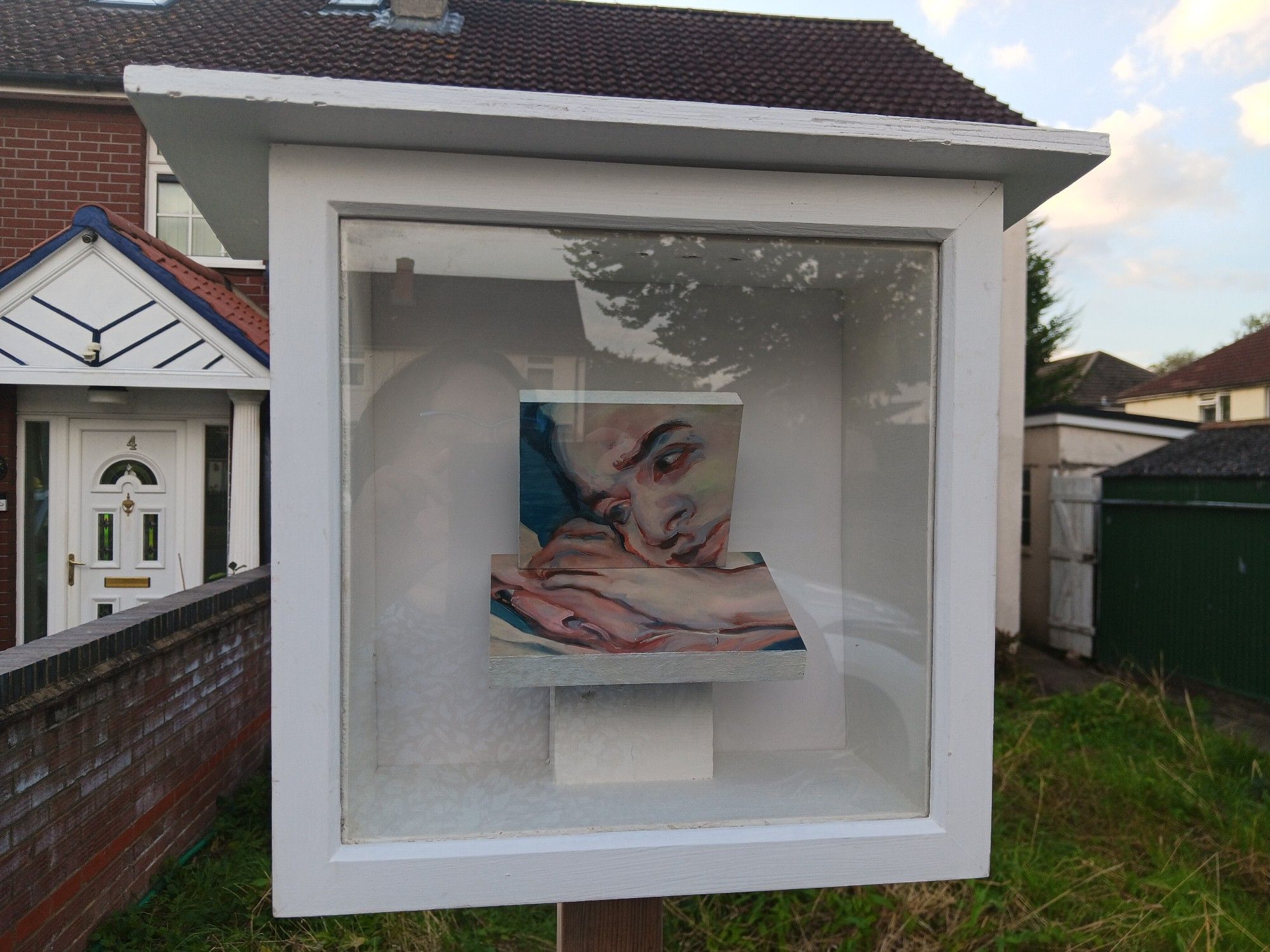 A square wooden white box with a glass front in a front garden. There is a very closed cropped painting of somebody with their head leaning on their hands inside.