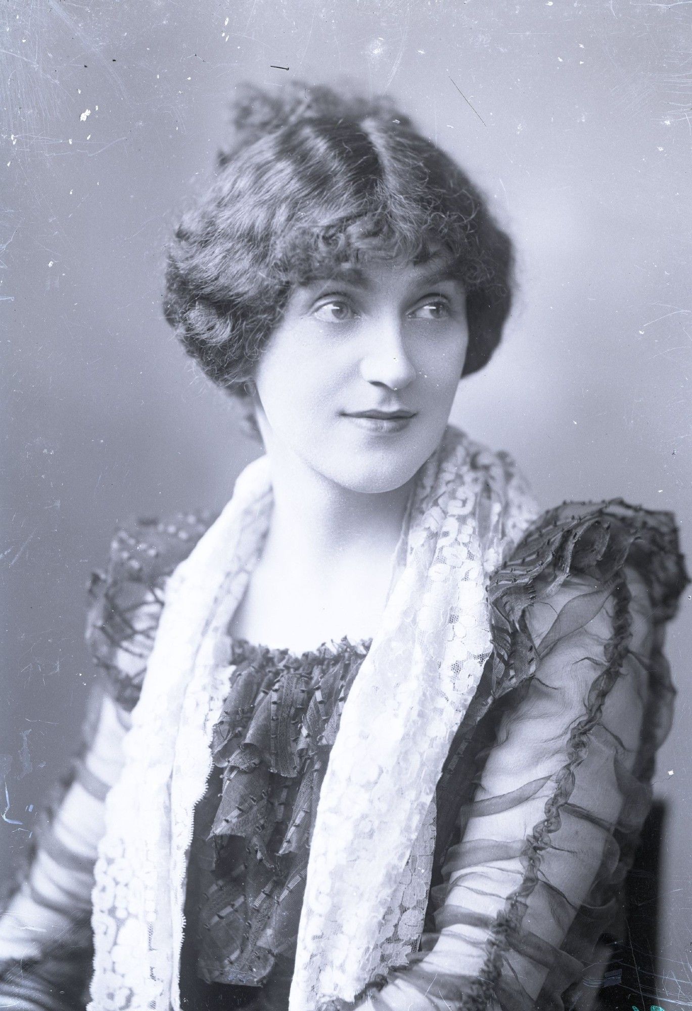 Diane de Rougy, a Victorian lady with dark brown hair in an up do, pictured sitting down from the waist up. She has a dress with see-through sleeves and a white scarf around her neck.