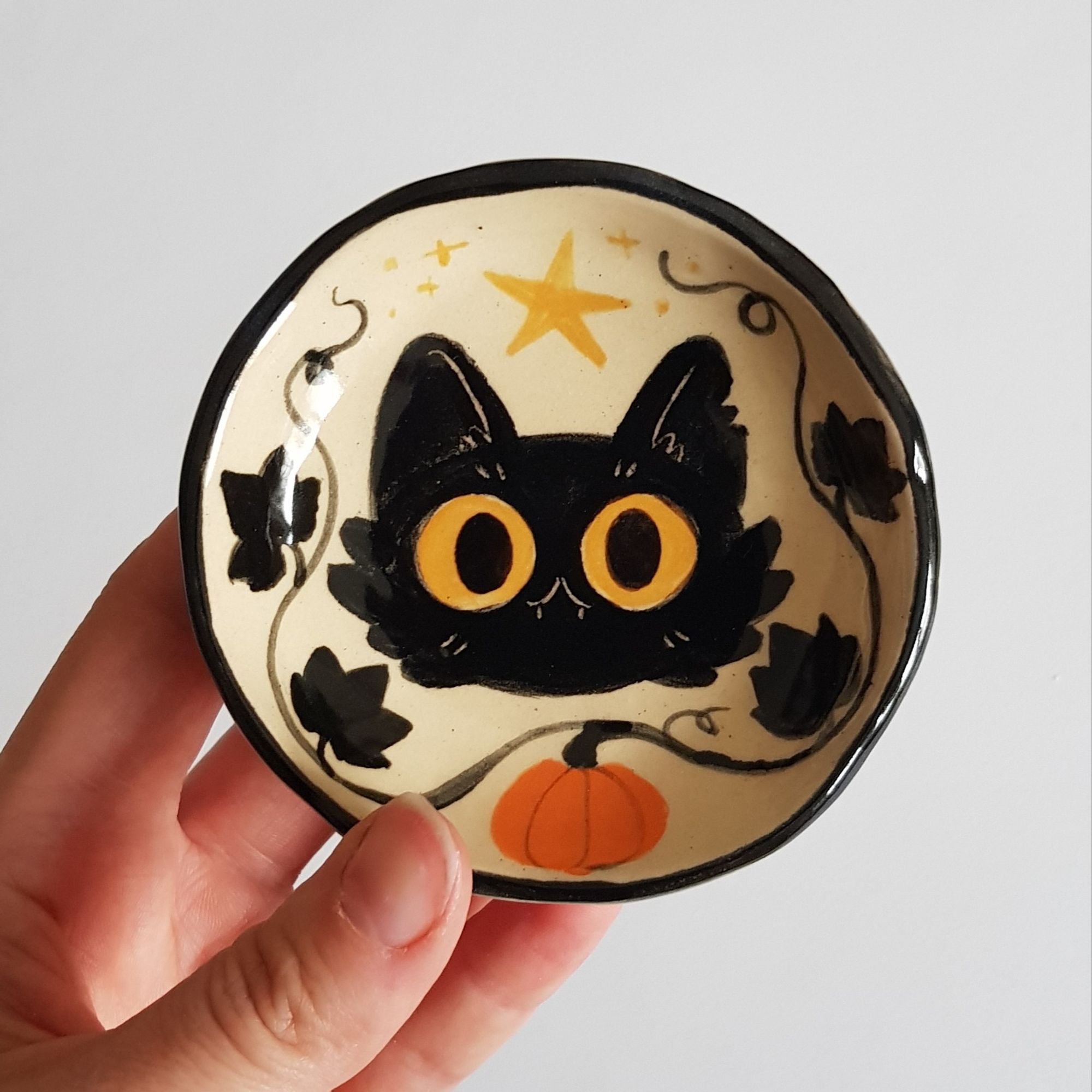 a photo of a hand holding a small ceramic dish painted to depict a head of a black cat with big yellow eyes and small fangs, surrounded by black pumpkin vines and a pumpkin at the bottom