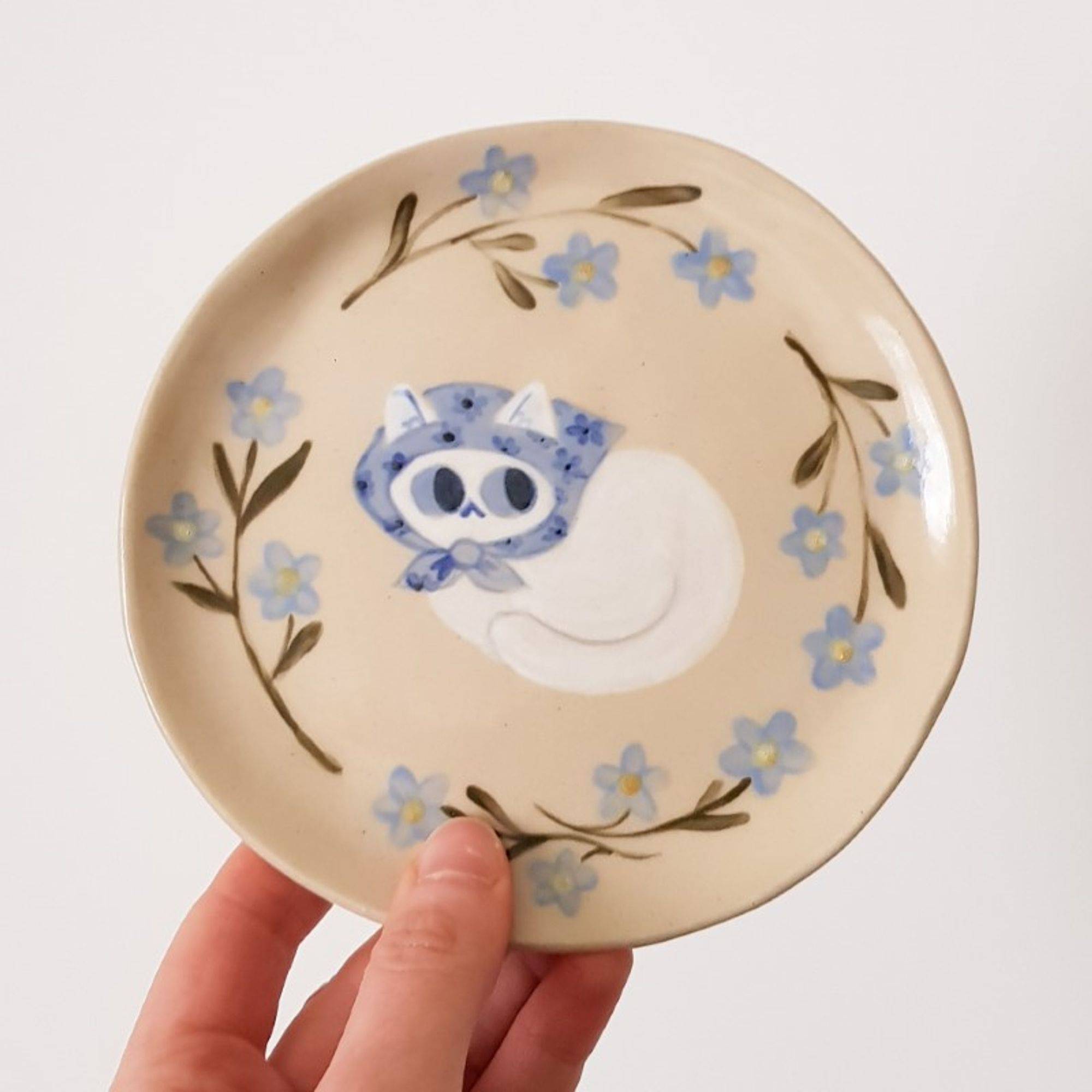 a painted ceramic plate depicting a white cat with a blue scarf on its head surrounded by blue flowers