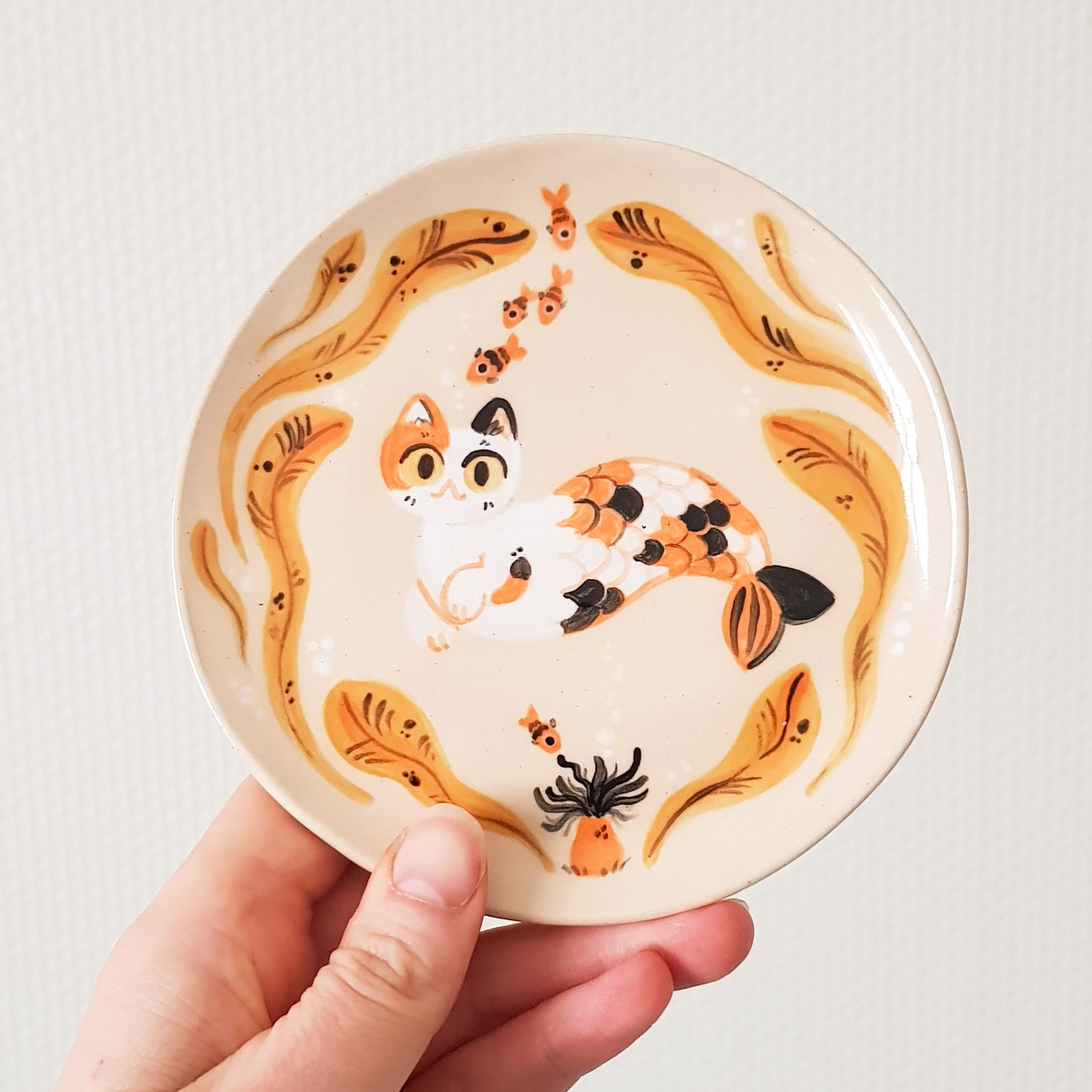 a small ceramic plate with a calico cat with a mermaid tail surrounded by yellow seaweed