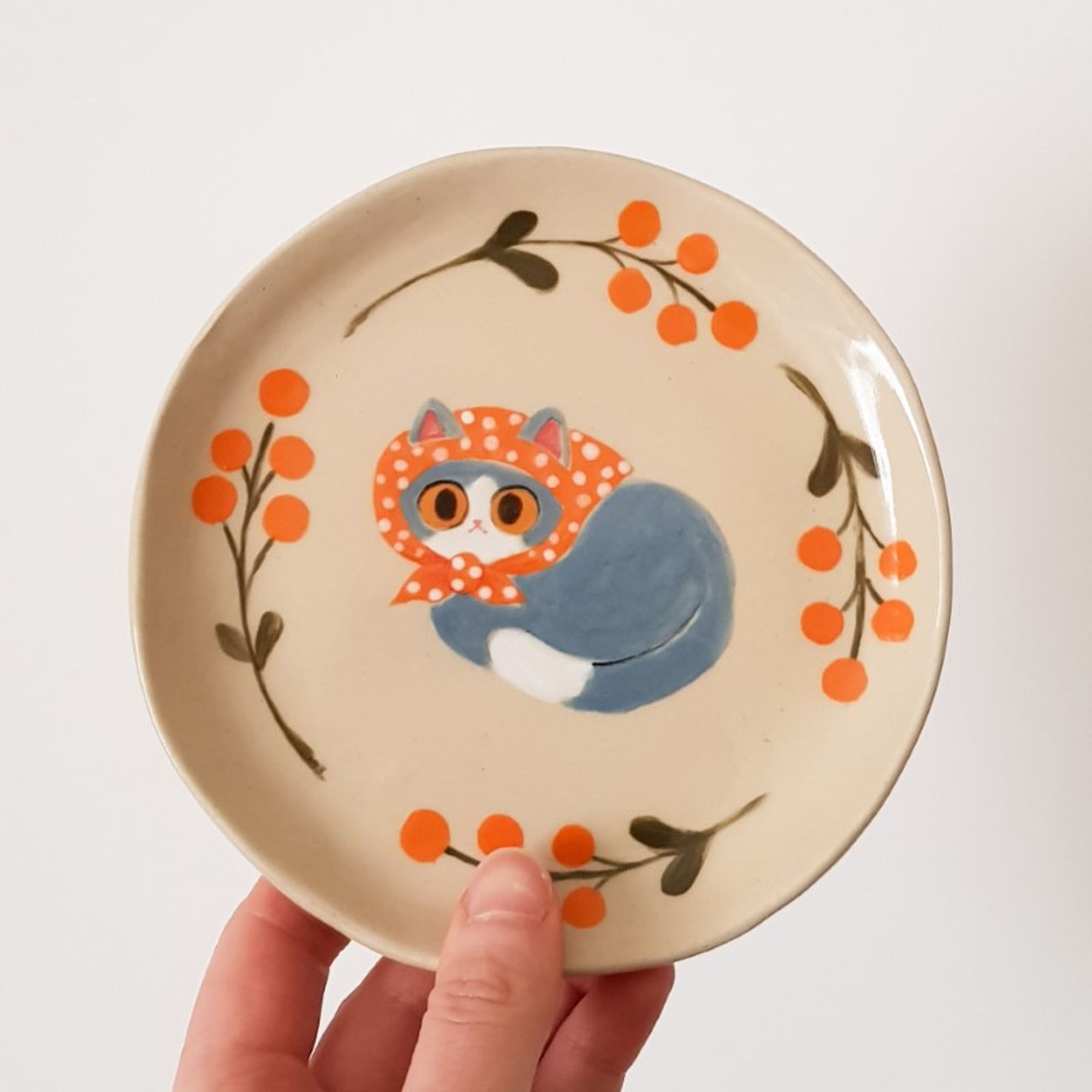 a painted ceramic plate depicting a grey tuxedo cat with an orange scarf on its head surrounded by orange flowers