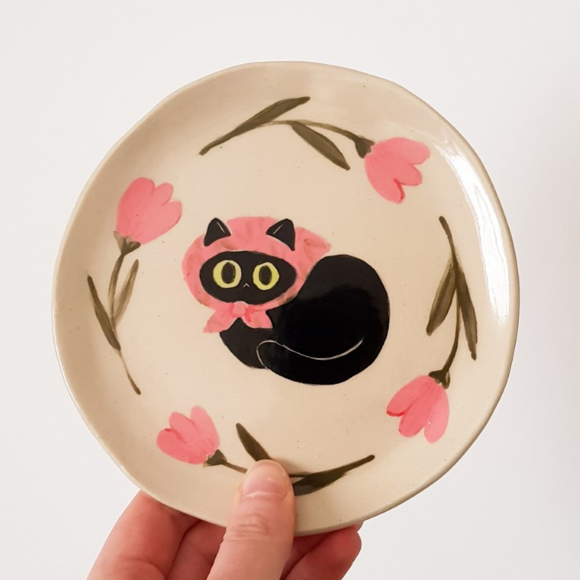 a painted ceramic plate depicting a black cat with an pink scarf on its head surrounded by pink flowers