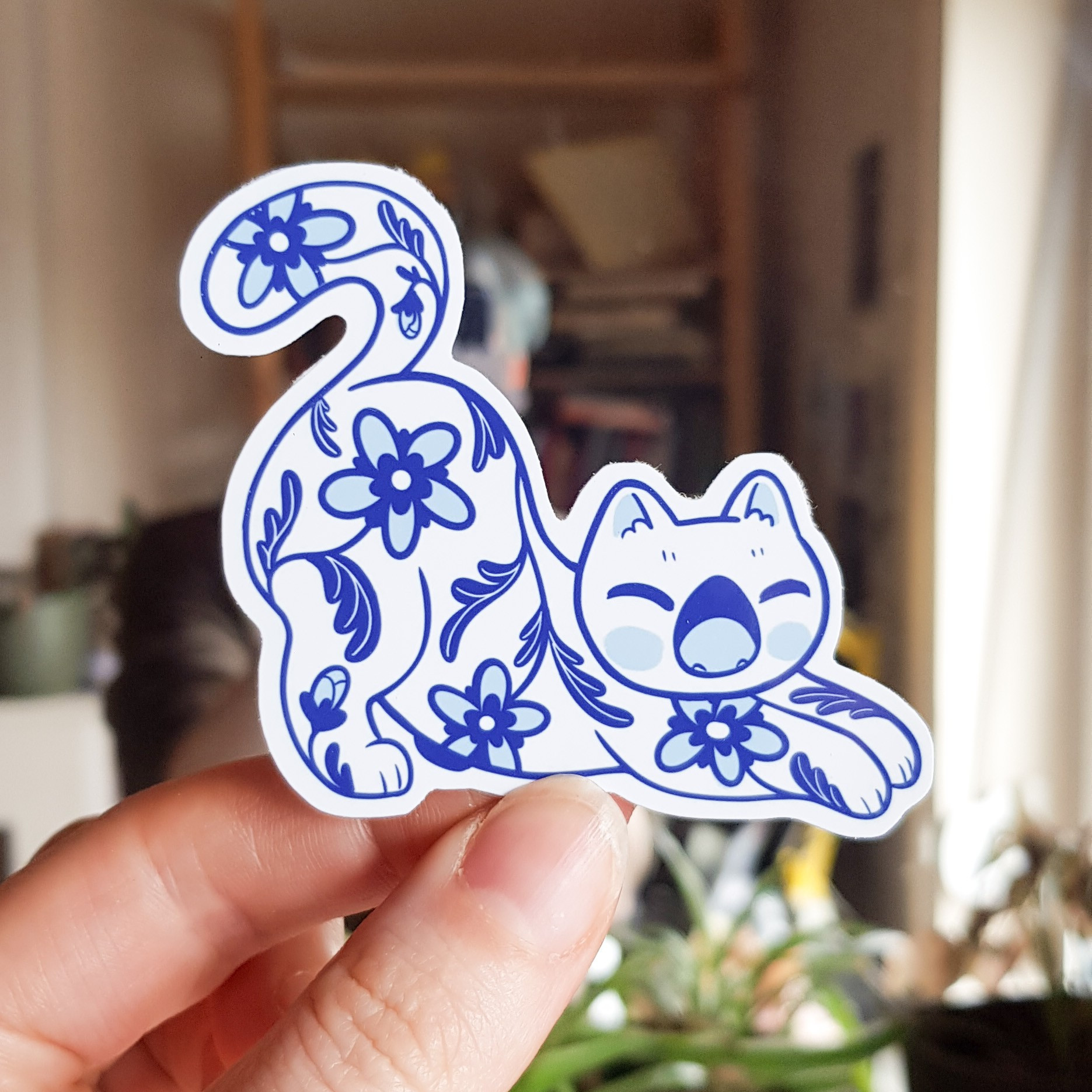 Photo of a sticker of a porcelain style white and blue cat decorated with flowers - cat is yawning and stretching