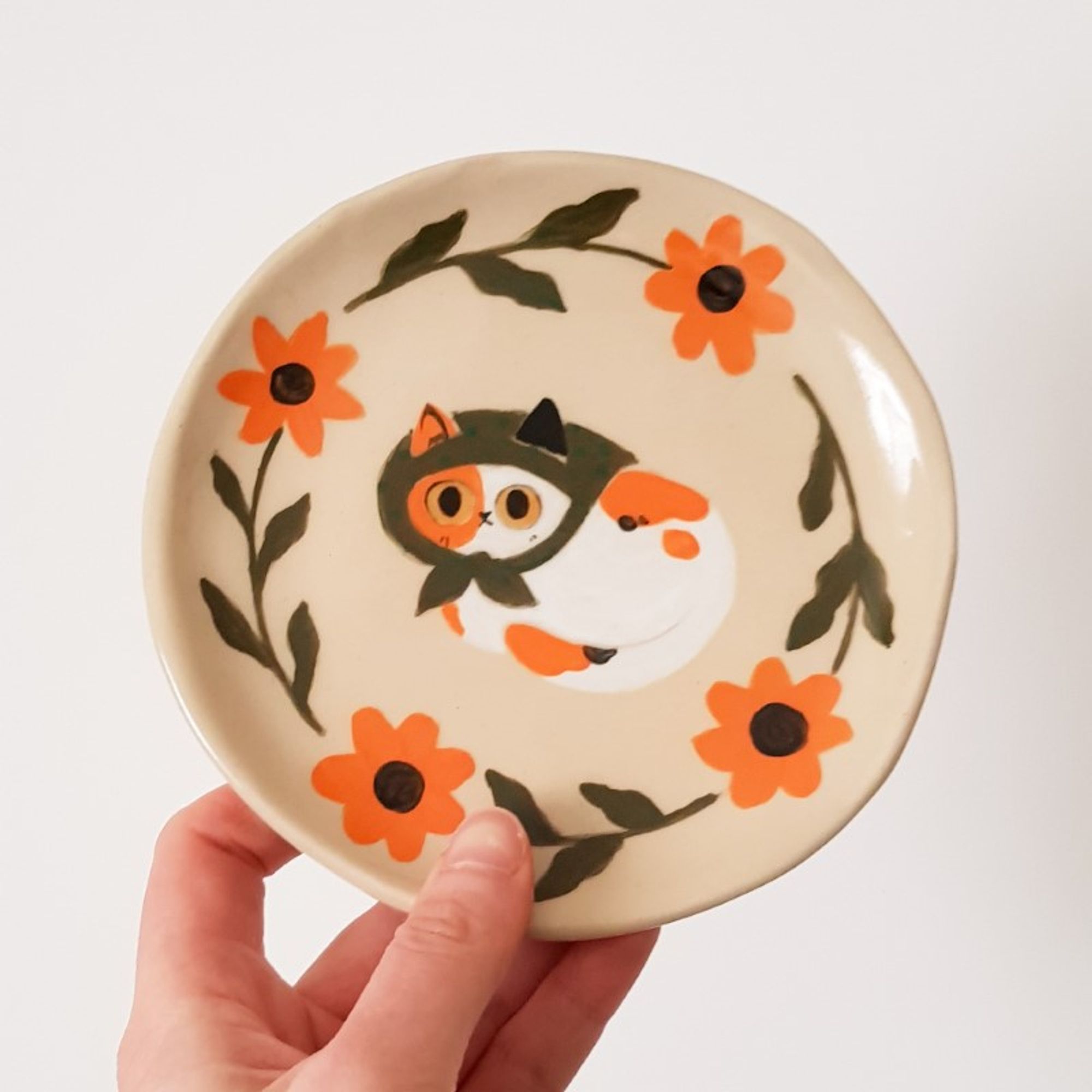 a painted ceramic plate depicting a calico cat with a green scarf on its head surrounded by orange flowers