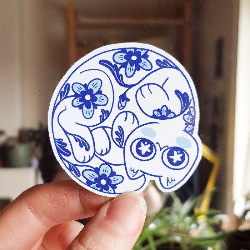 Photo of a sticker of a porcelain style white and blue cat decorated with flowers - cat is rolled up in a circle, looking up