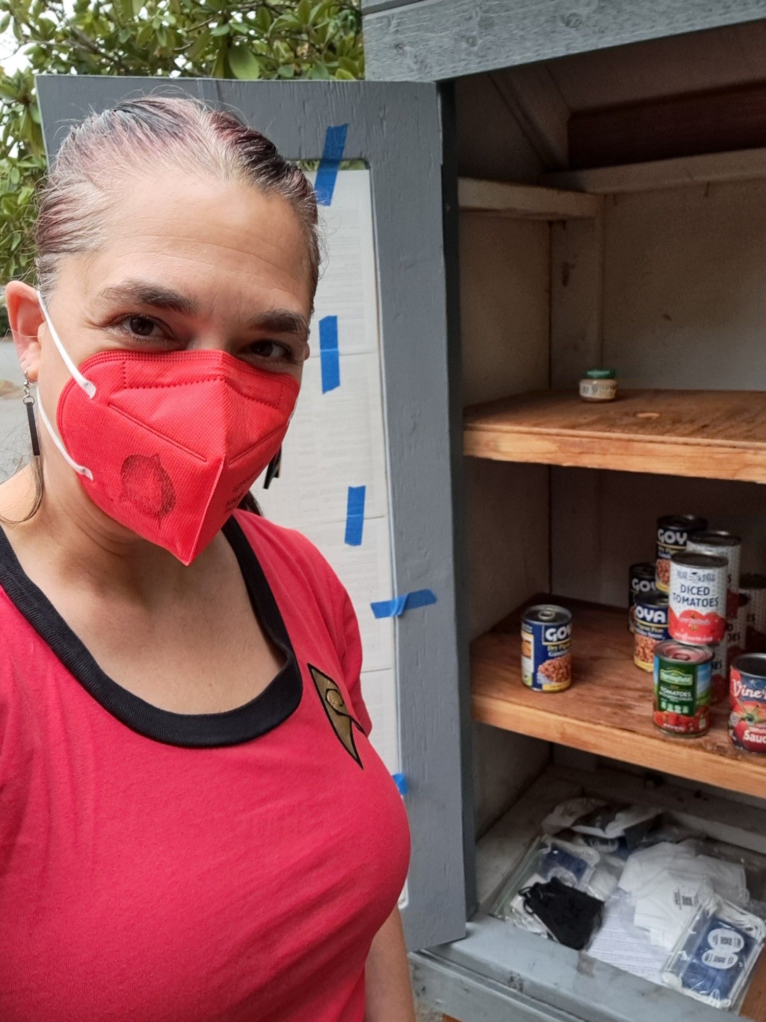 A white woman with brown hair and eyes wearing a red Original Series Star Trek T-shirt dress, rainbow Starfleet delta earrings, and a red Powecom KN95 with a Starfleet insignia stamped on it in black. She is standing in front of a Little Free Pantry.  The bottom shelf has an assortment of individually wrapped masked and the middle shelf has canned vegetables.