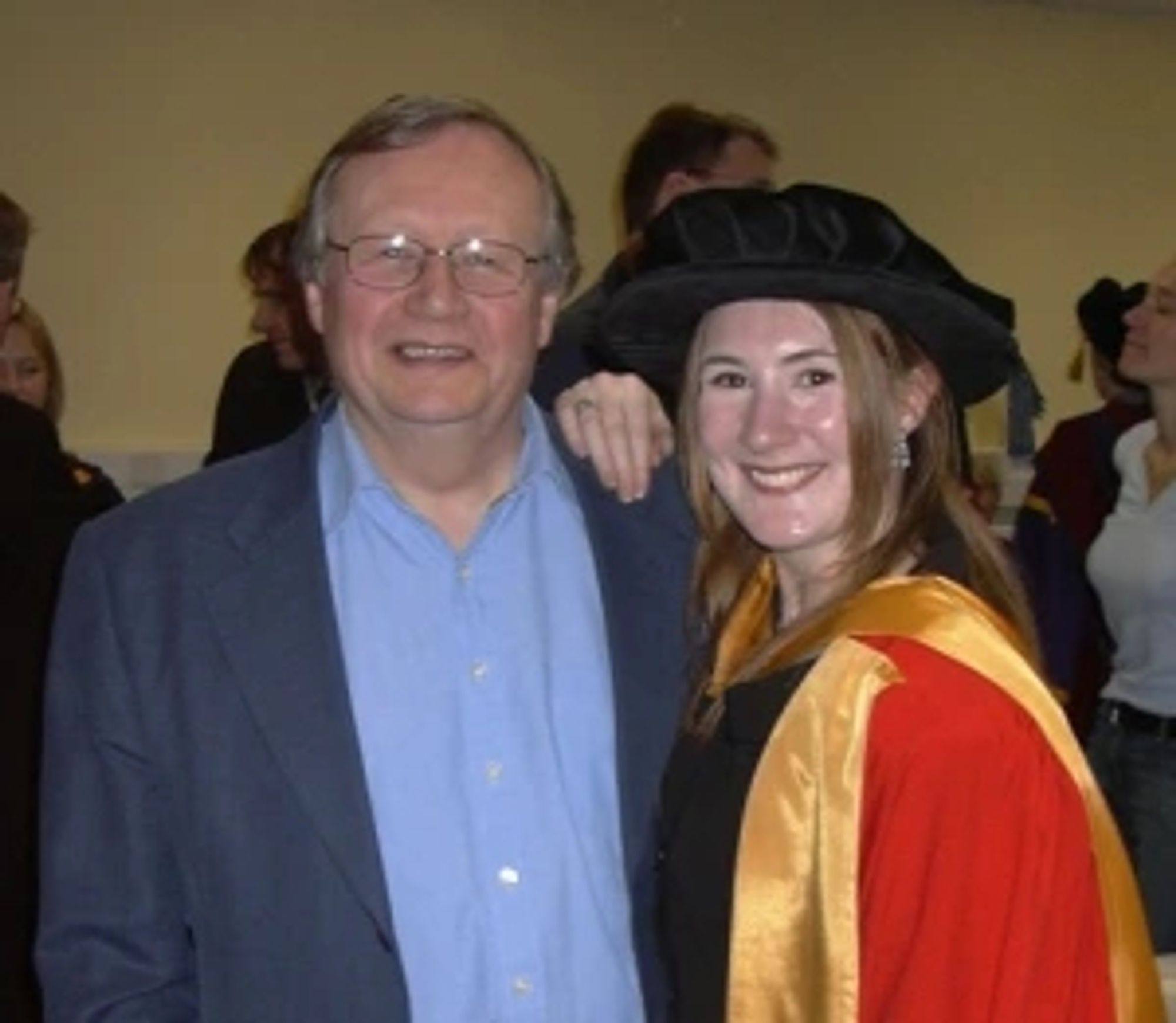 Photo of a man with glasses wearing a blue shirt and jacket stood next to a woman wearing a red and gold graduation gown and black  hat