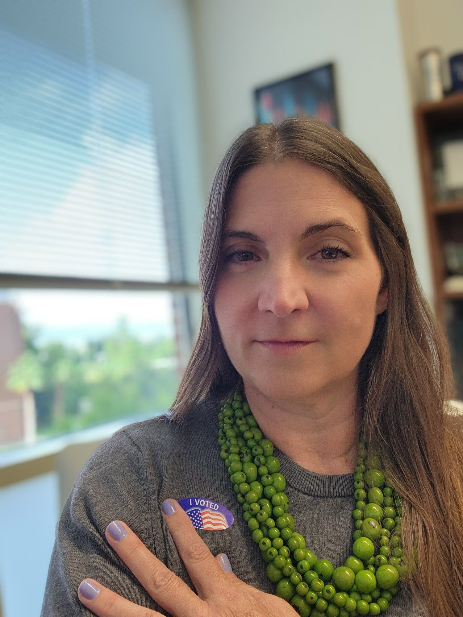 Middle-aged female professor with brown hair and bold green necklace pointing to "I voted" sticker on her gray sweater with lavender-colored fingernails.