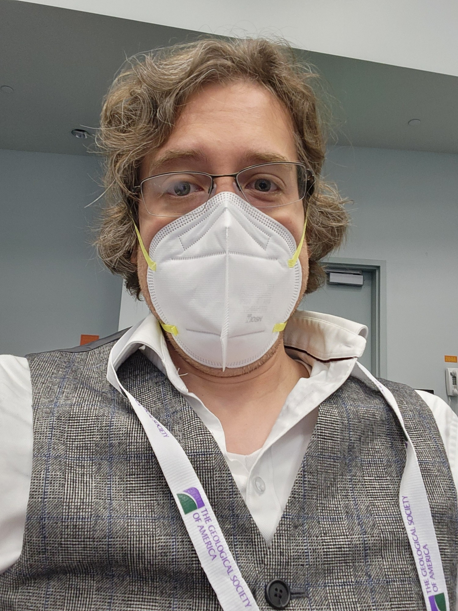 A photograph of me, a white man with midl-length wavey hair and glasses, wearing a grey waistcoat over a white shirt and a facemask.