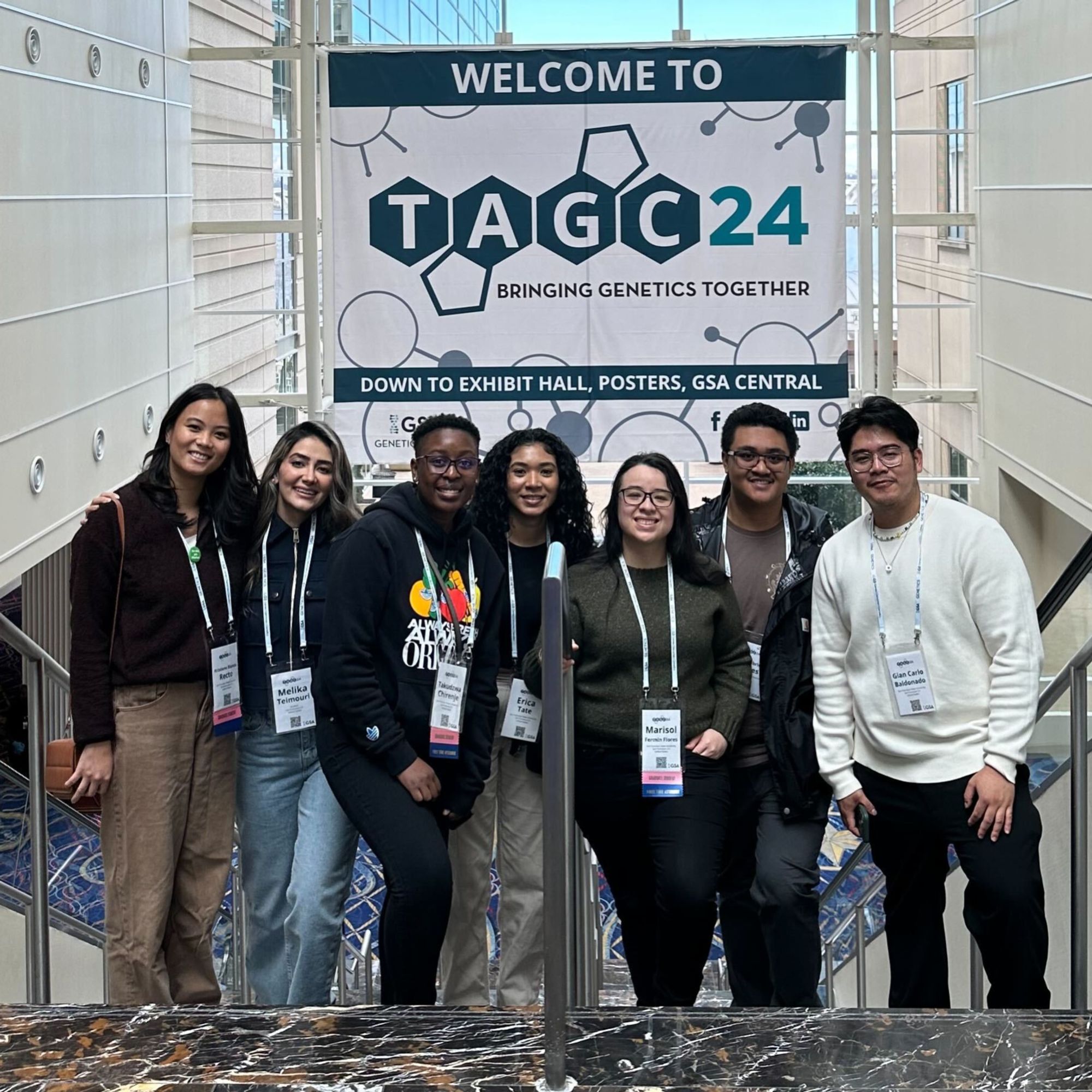 Seven students from San Francisco State University with a sign that says "TAGC 24 bringing genetics together" and "Down to eshibit hall, posters, GSA central"