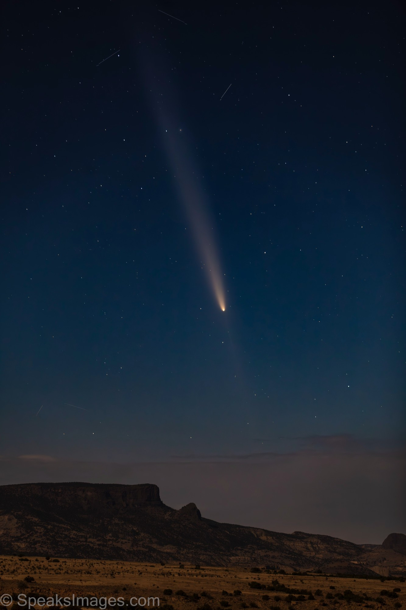 Astro-landscape photo of comet with long tail above moonlit mesas. Anti-tail is faintly visible below coma of comet. 