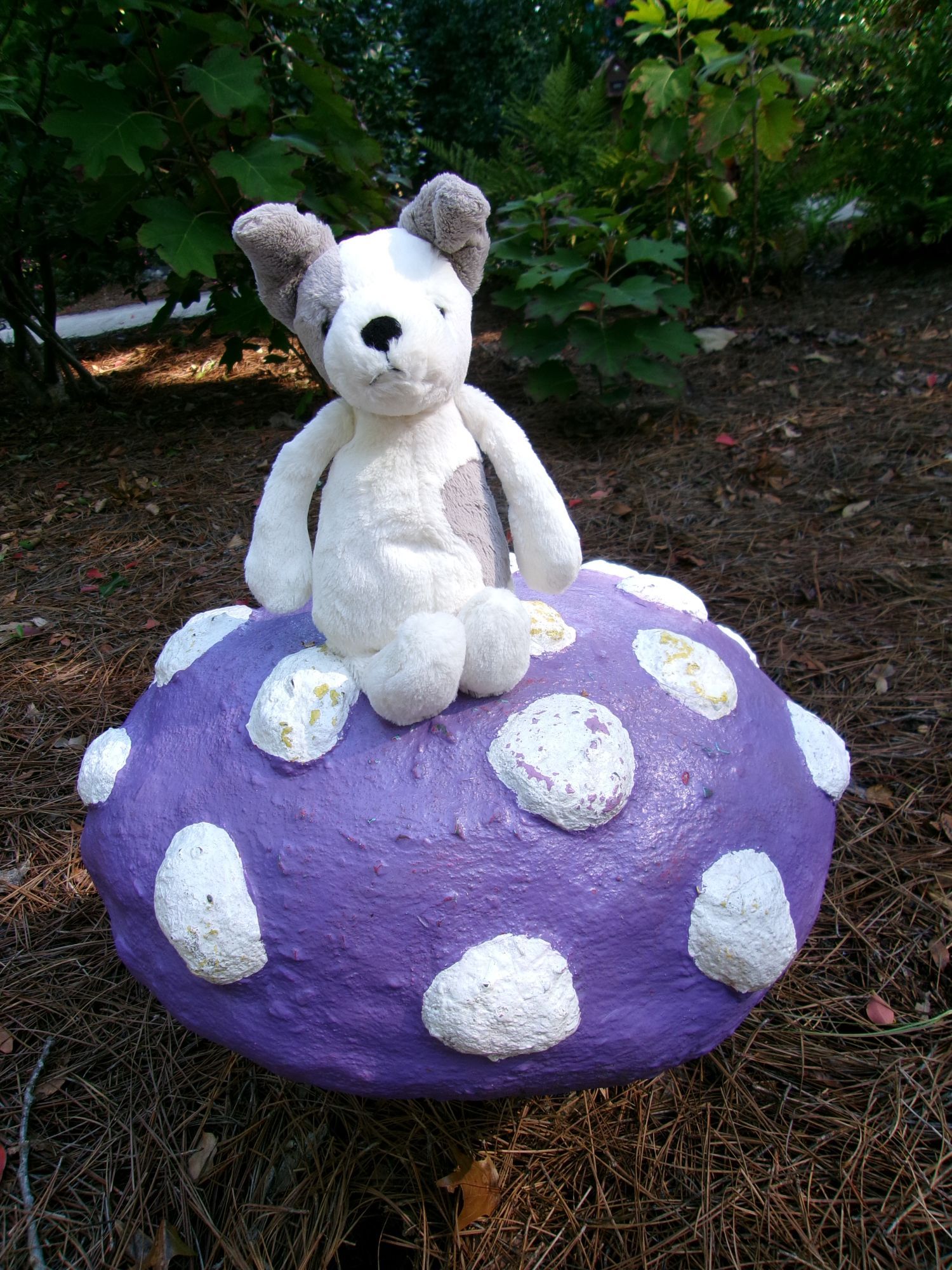 A photo of a white and gray dog plushie sitting on a purple toadstool statue in a park.