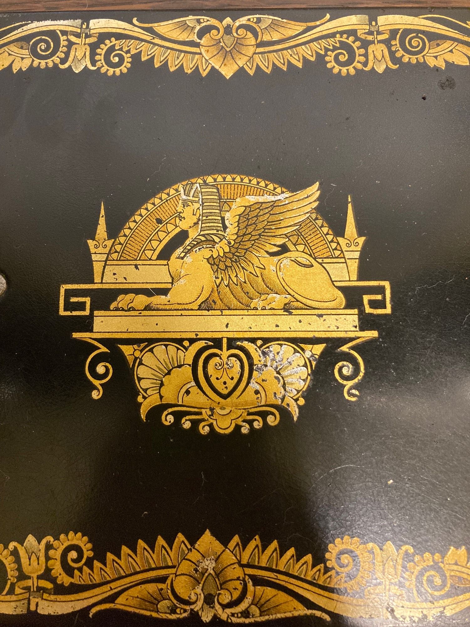 A close-up of some of the aforementioned decoration: on a black lacquer background, a gold Sphinx is shown in profile in the middle of the machine’s bed with some vaguely fern-like scrollwork above and below. The gold is chipped from many years of use.