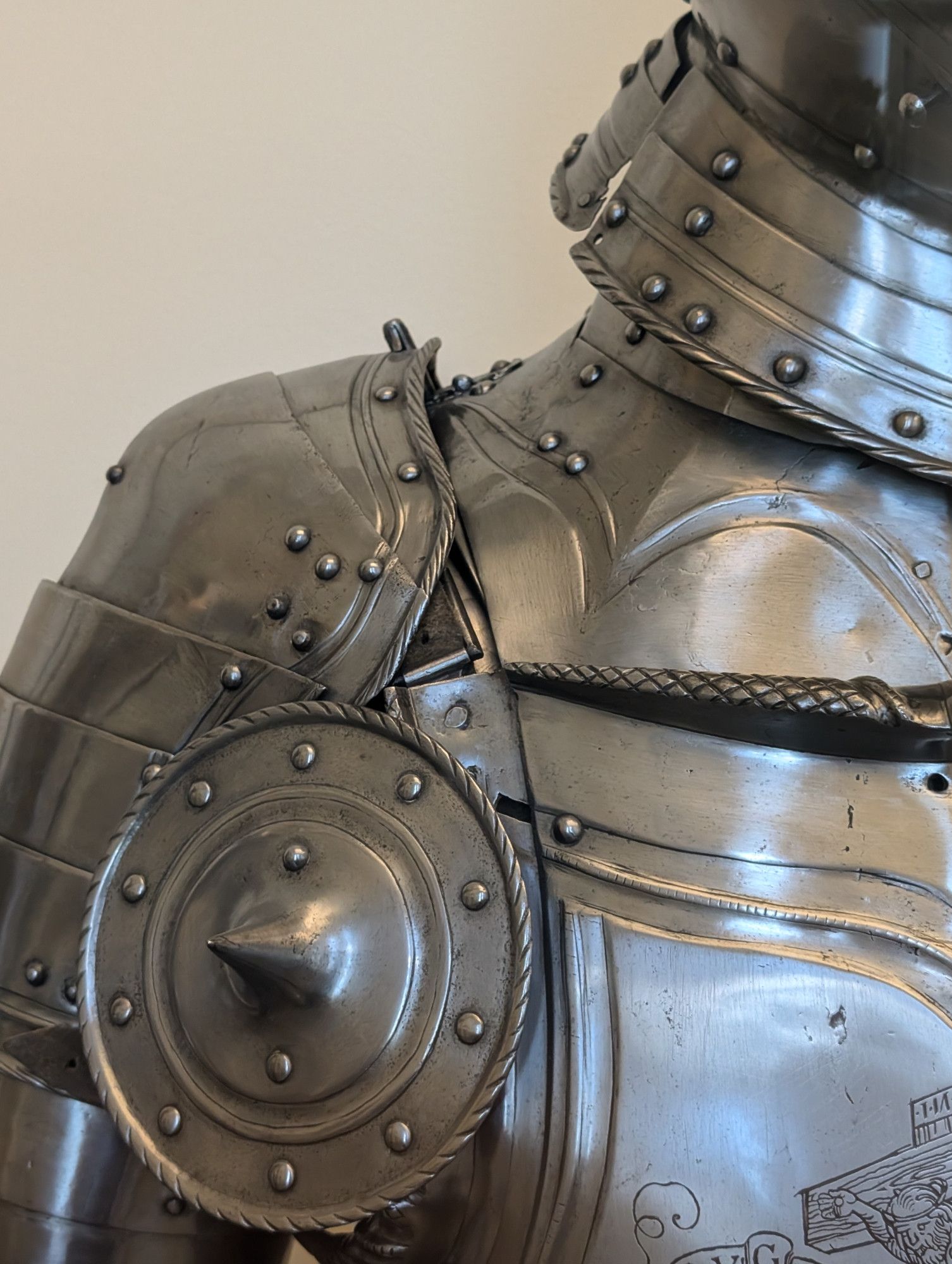 A closeup of the shoulder intersection of the cuirass, pauldron, and bevor of a piece of 1500s field armor. Some ornate etching of the crucifixion is visible in the lower right corner on the cuirass.