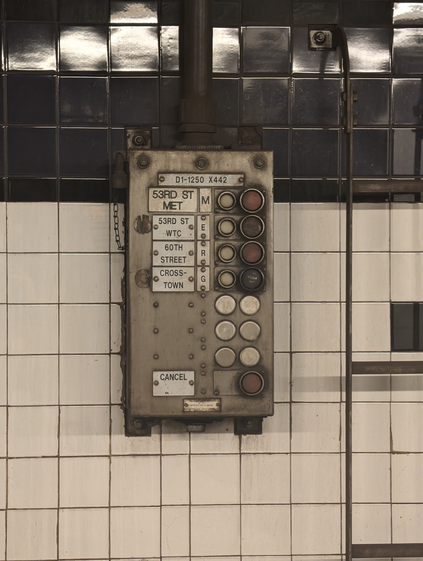 Some kind of switch board on the NYC subway. It has big red buttons, labels of stops, and it's coated in grime. It hangs on a shiny black and white tiled wall next to a metal ladder.