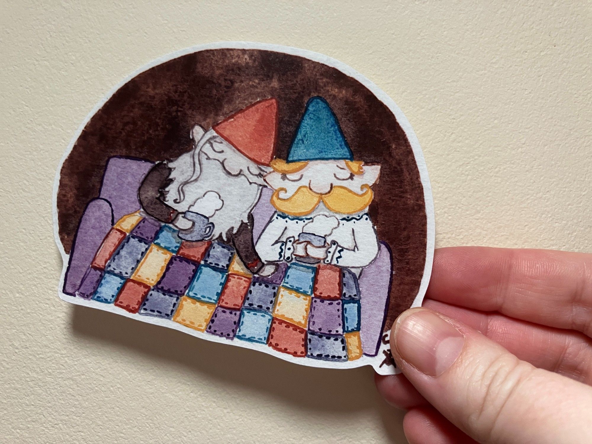 A sticker of two gnomes snuggled down together beneath a patchwork blanket.