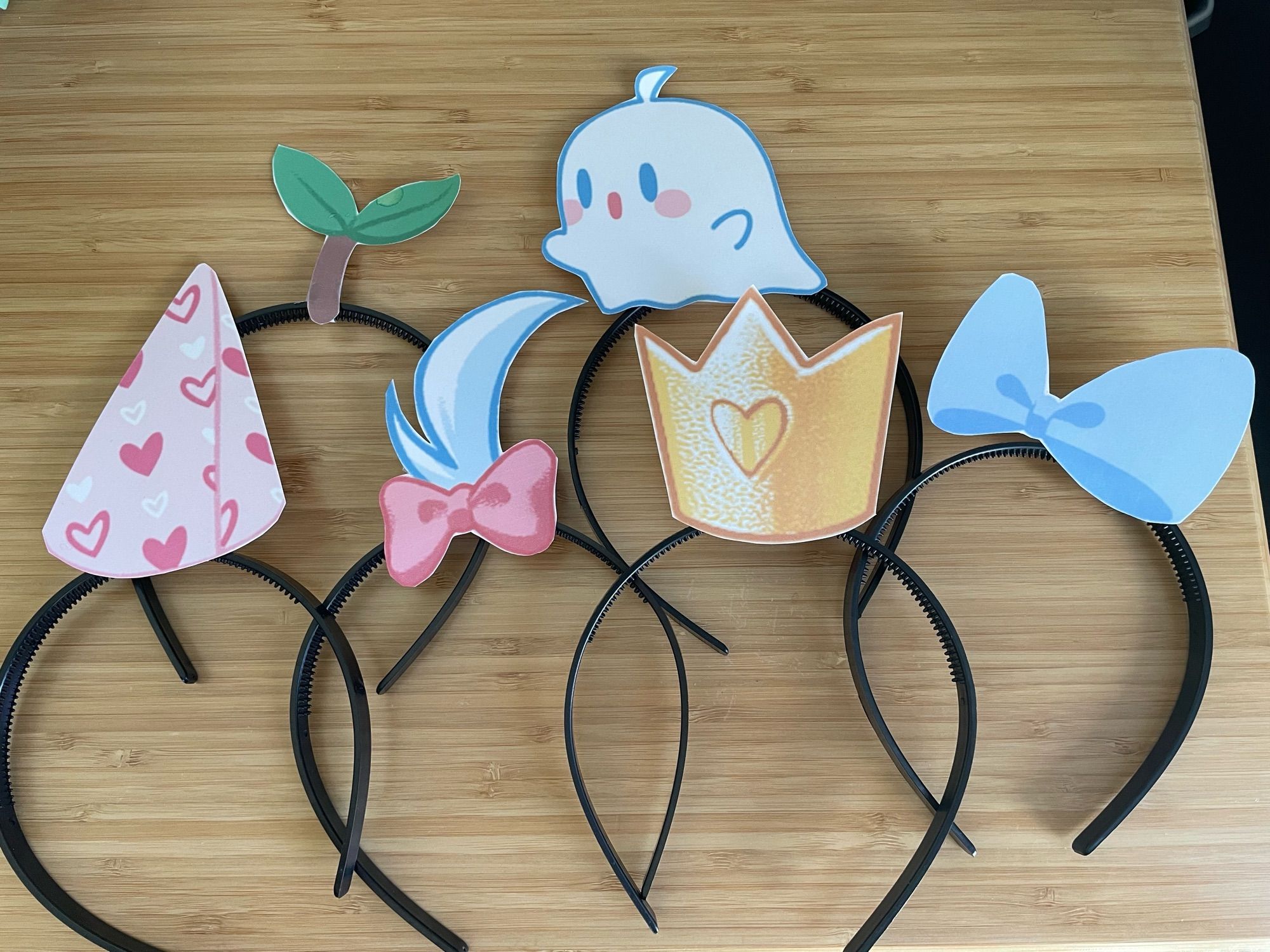 6 headbands laid out on a table, each one has a different “hat” attached, like a crown, bow, mini ghost, party hat, Mina’s hair and bow, and a plant sprout