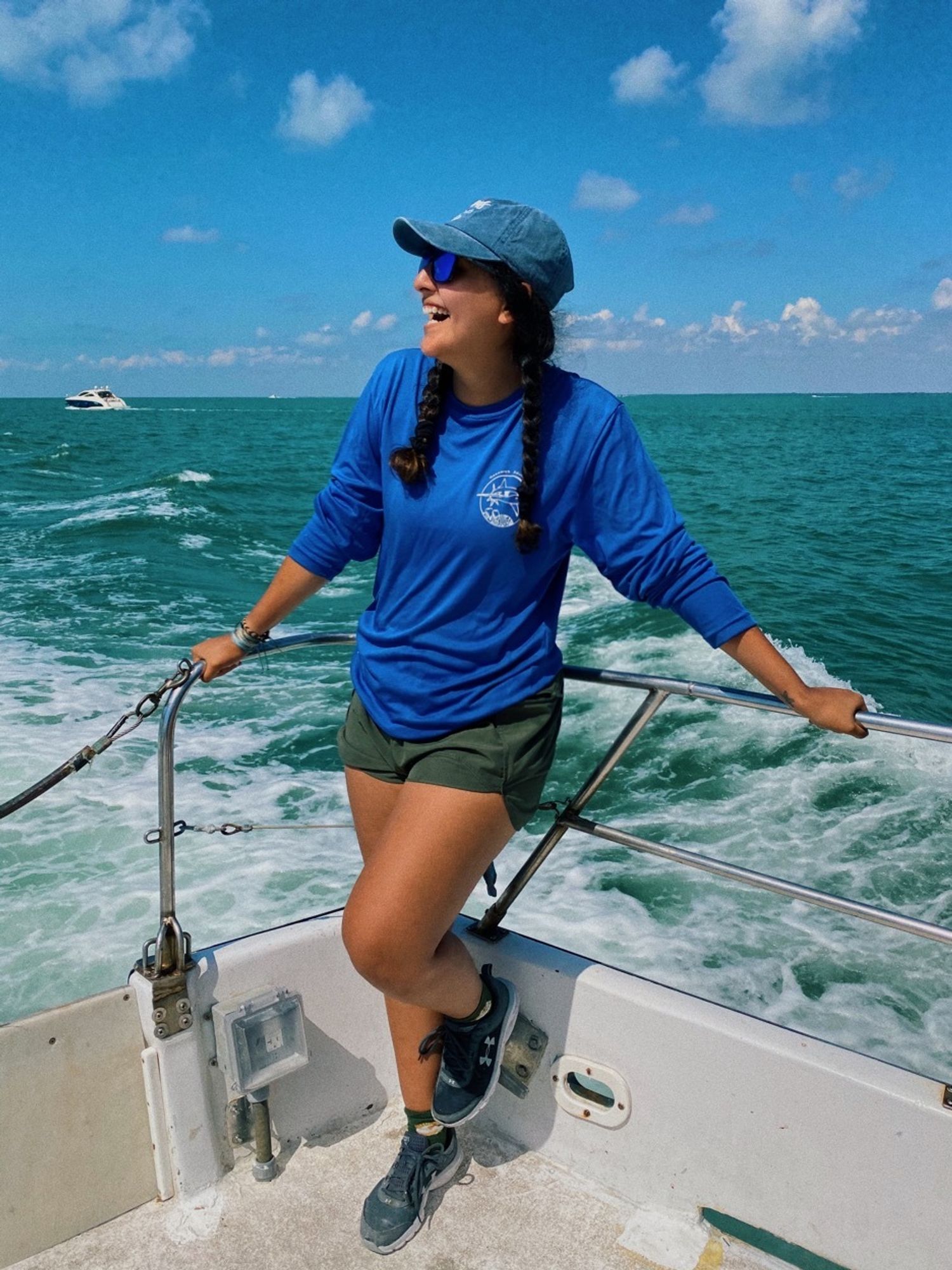 I am standing on the railing of a moving boat off the coast of Miami, the water is very blue/blue green, i am holding onto the railing and i am smiling to the left. I am wearing greens shorts, grey shoes, and a blue longsleeve as well as a blue baseball cap on. My hair is in pigtail braids and i am wearing sunglasses. It is a partly cloudy sky