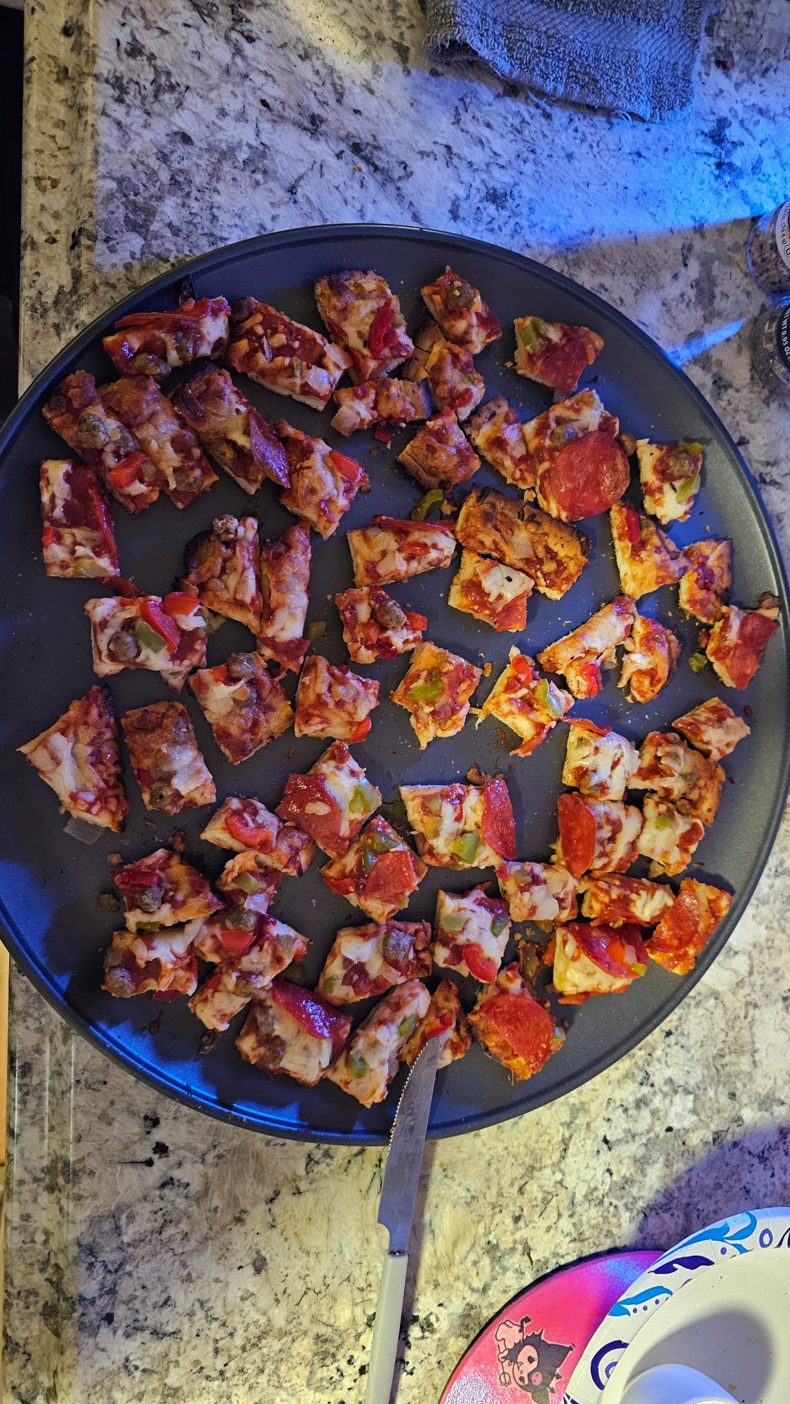 A supreme pizza, cut into semi-regular sized pieces, sits on a pizza pan on a faux-granite counter top. 