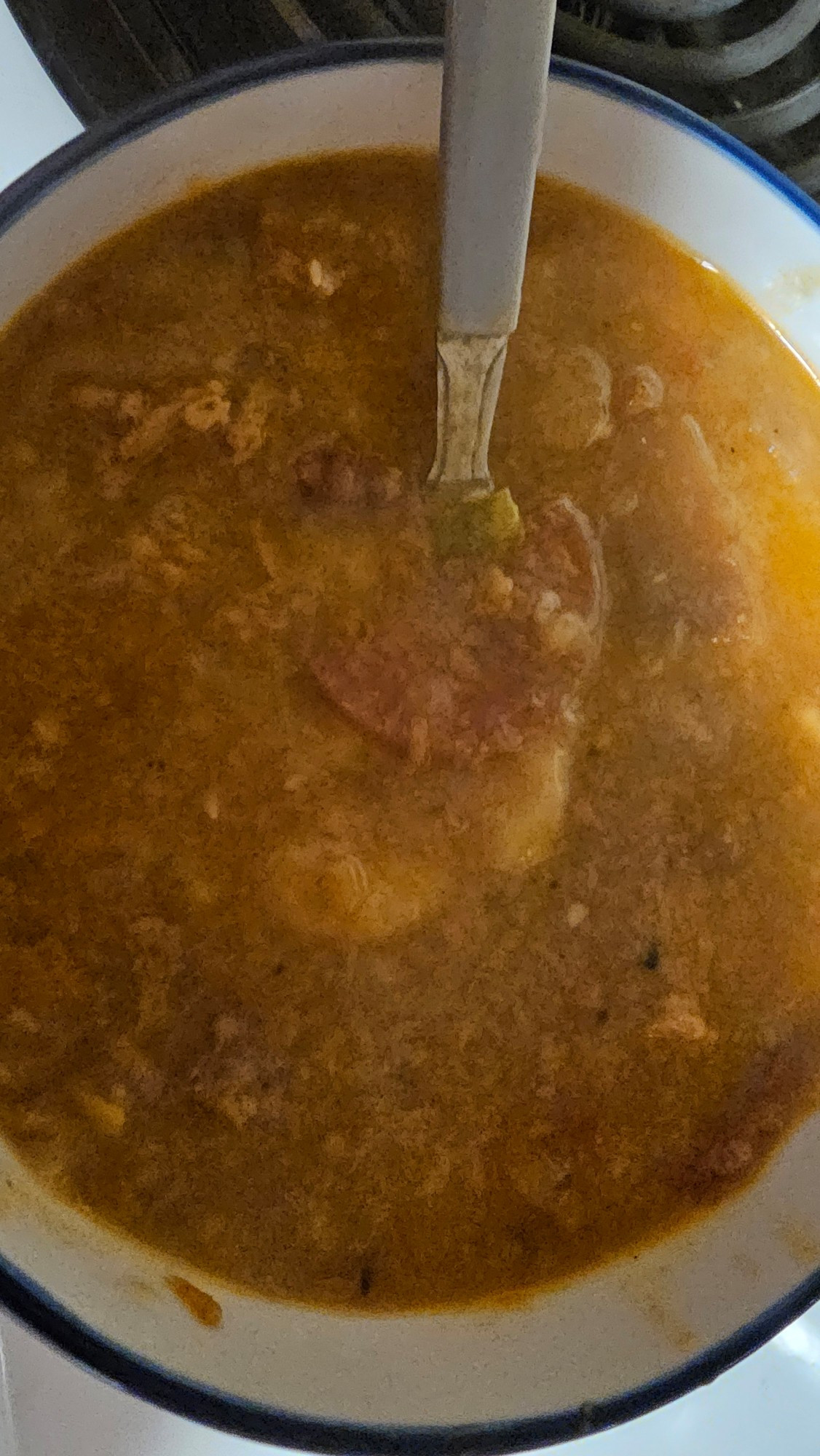 A bowl of Pizza Soup sits on a stove top with a spoon resting inside. There is a piece of pepperoni visible.