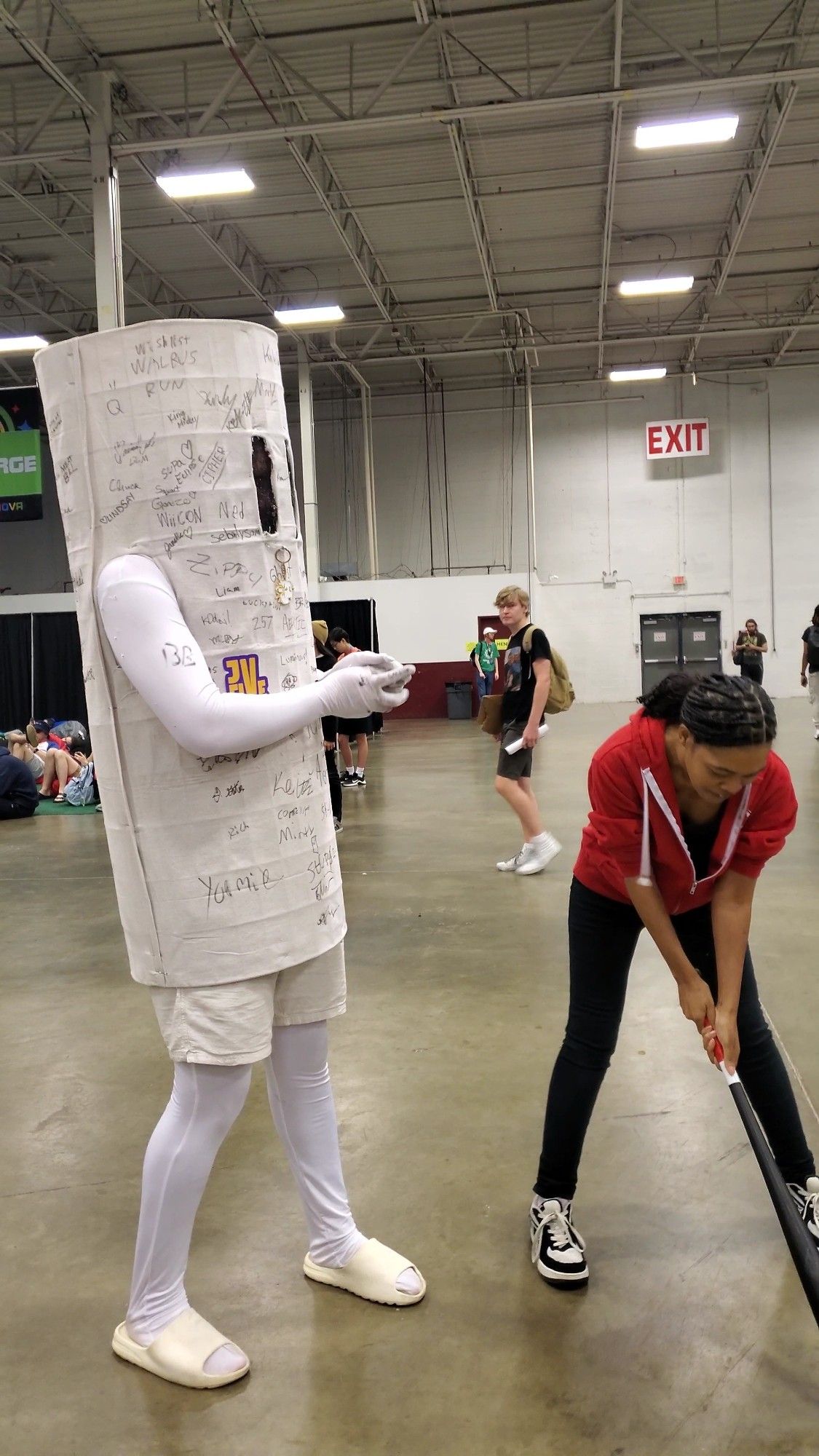 Its a picture taken from the Dulles Expo Center in Chantilly, Virginia during SuperNOVA. Sharpie is wearing a red zipup hoodie with black pants and is holding a baseball bat. To the left of her, a person is cosplaying as the smash bag from Super Smash Brothers.