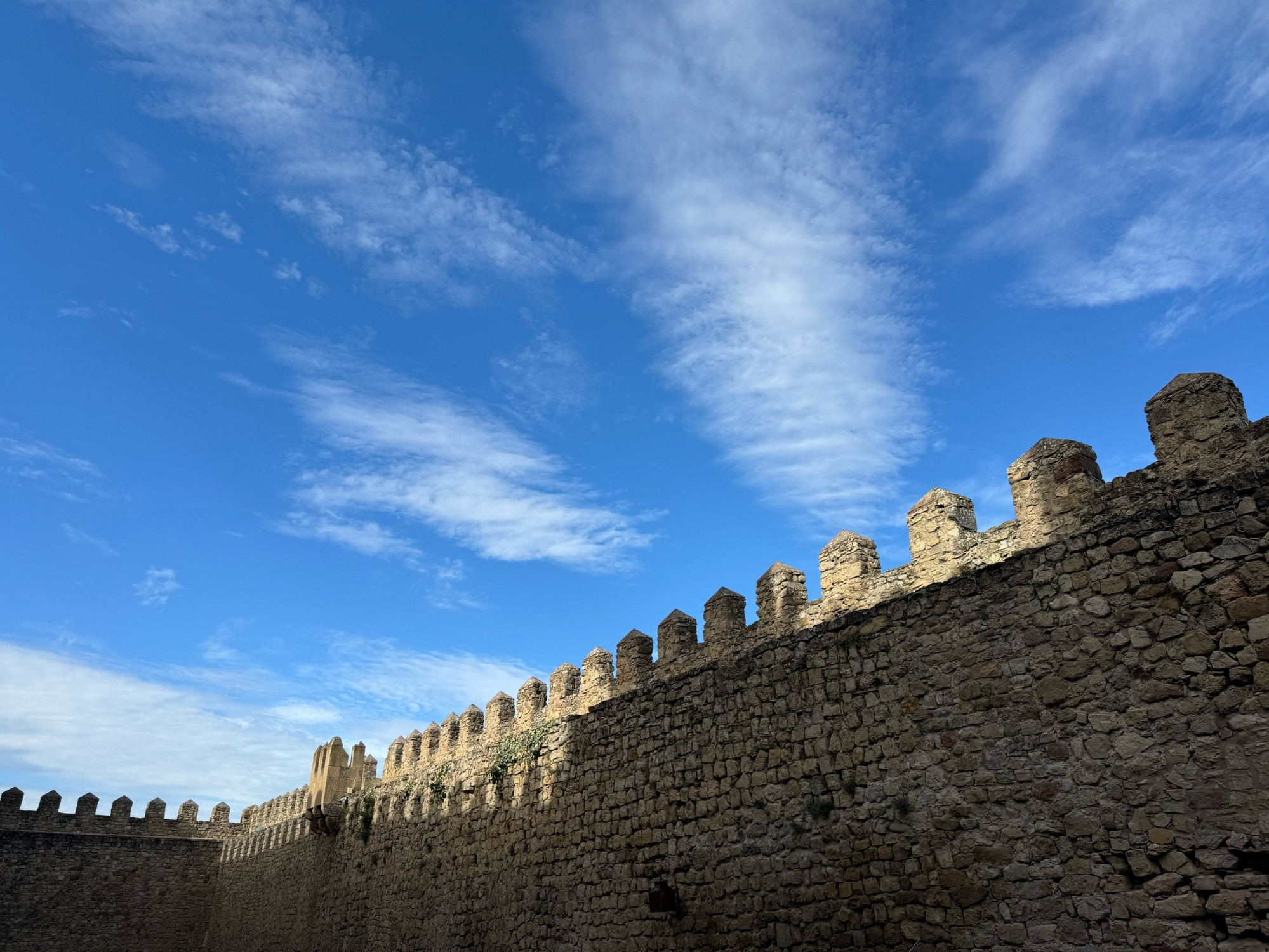 photo of the top of a castle wall made of stone. above it the sky is blue with small puffy clouds. 