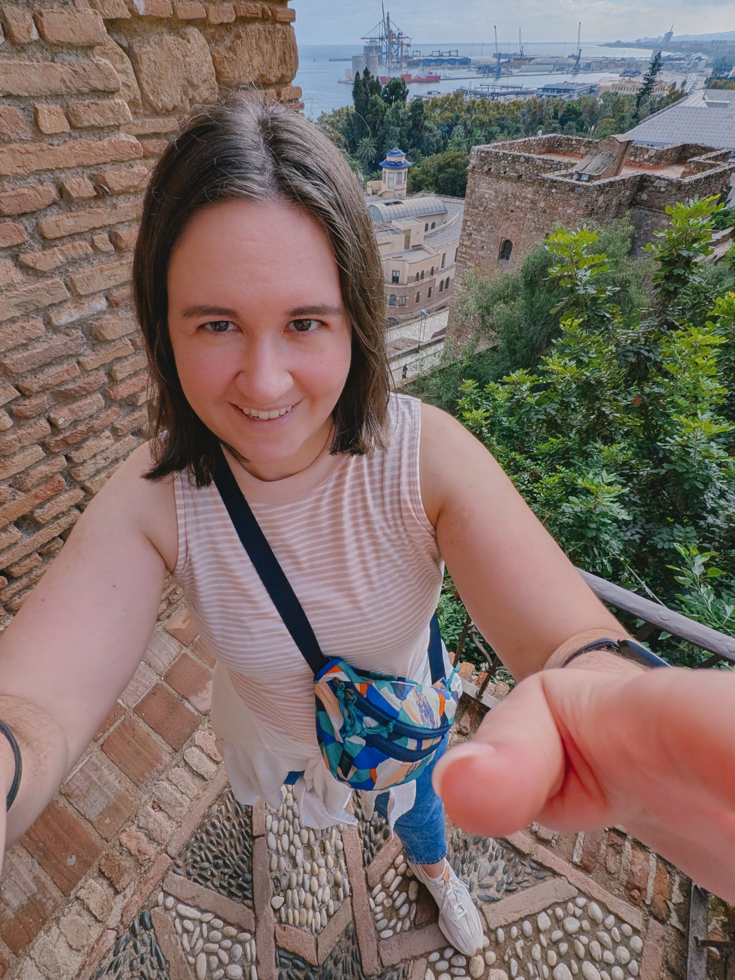 photo of Bella taken from above with a slight fish eye lens effect. Bella is looking up and reaching up for the camera. behind them is a brick wall and then a high up view of part of a city with lots of trees and an ocean port area. 