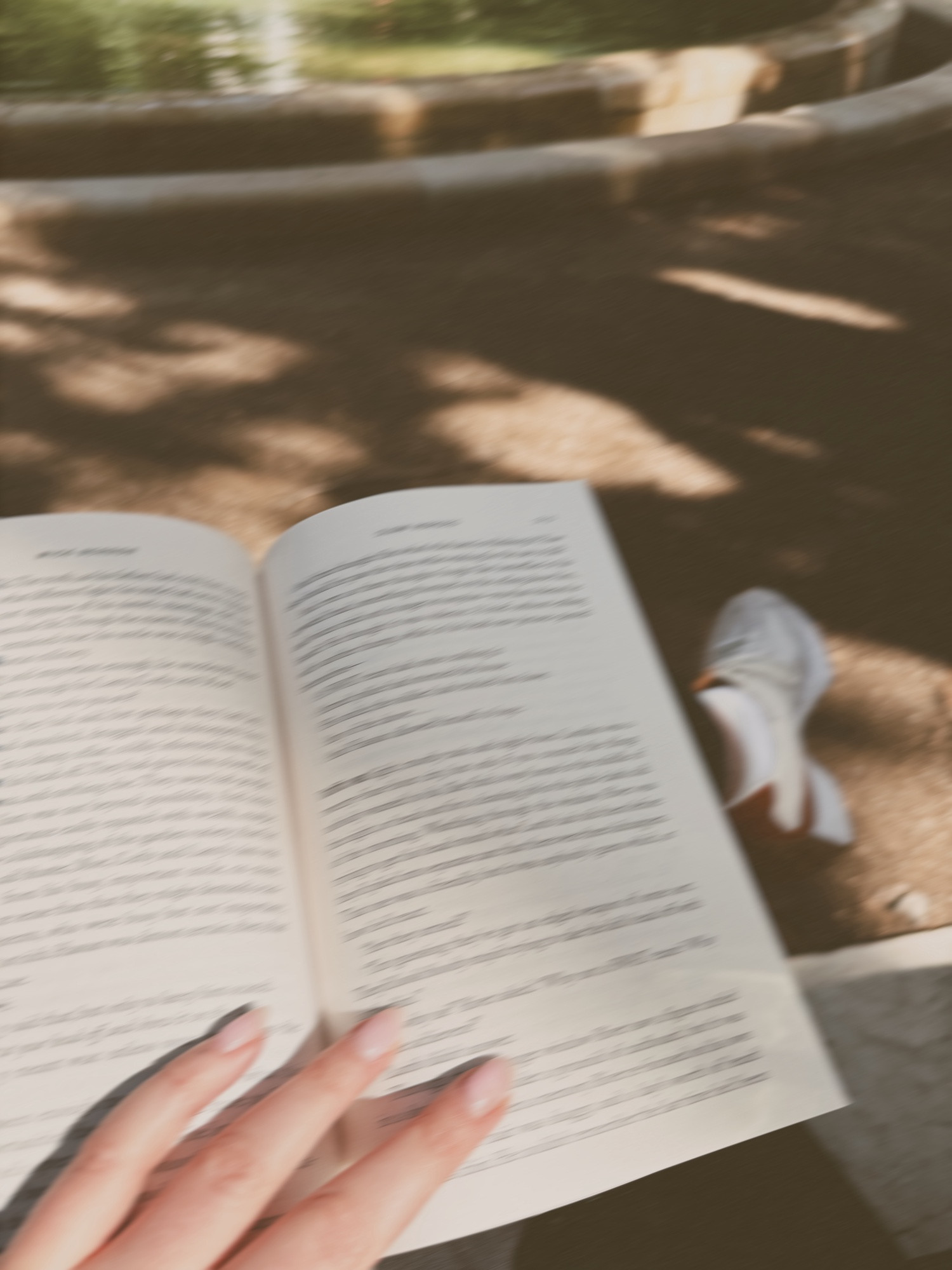 Photo of Bella’s hand holding open a book resting on their lap. Their shoe is peeling out behind the book and the background shows a dirt trail with shadows of tree leaves and a small bit of a fountain. The photo is slightly blurry. 