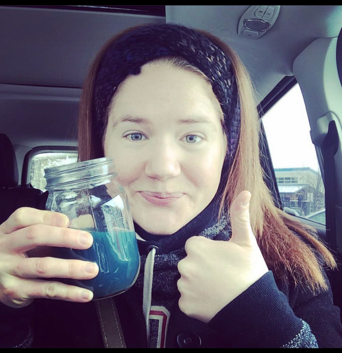 Selfie of Kelly giving a thumbs up in her car while holding a mason jar full of bright blue liquid (spirulina mixed with lemon and Himalayan salt). She’s wearing a roots sweater and headband.