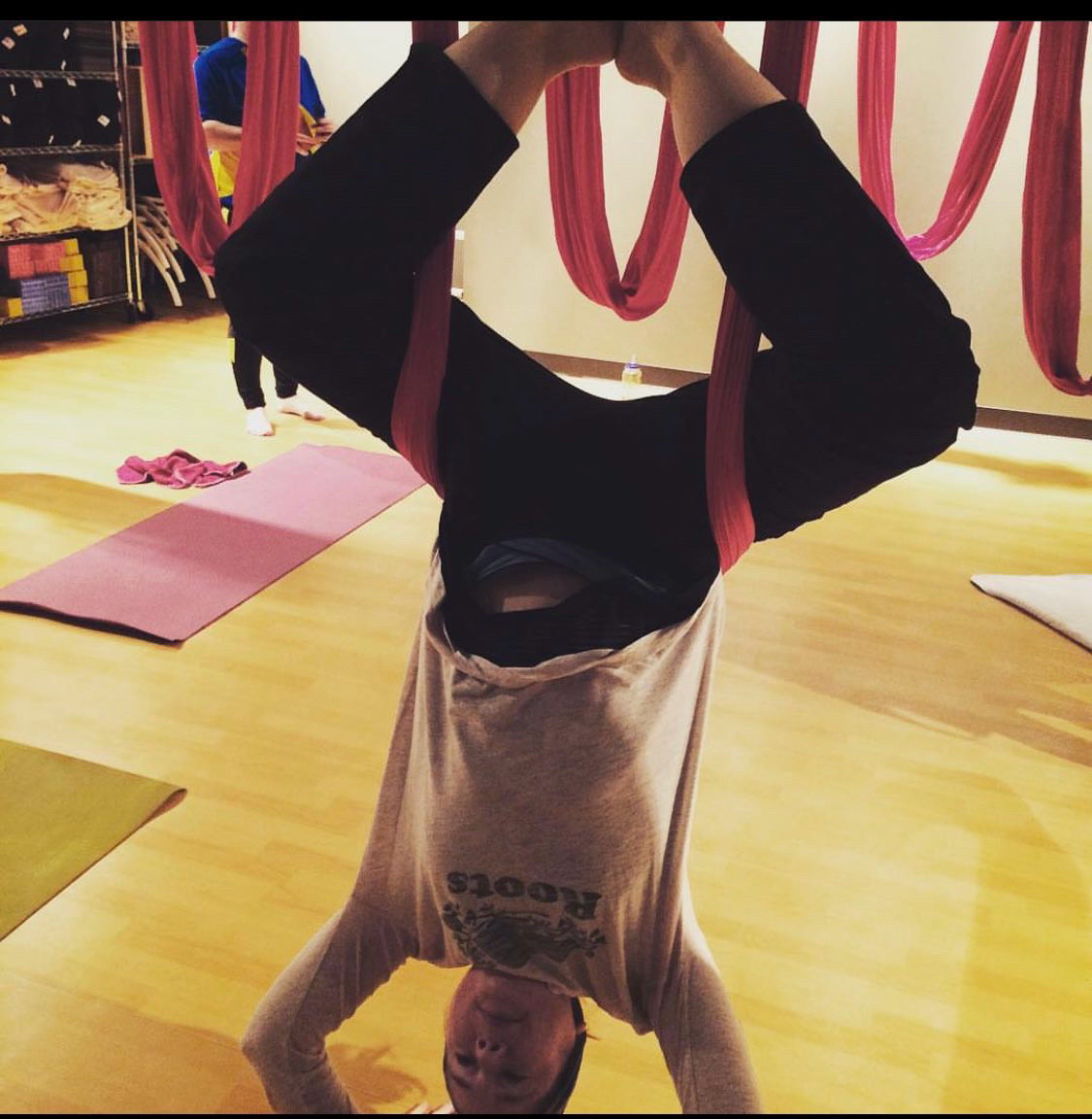 Kelly hangs upside down suspended with red aerial yoga ribbons. Her arms are on the floor in a handstand pose and she’s wearing a white roots tshirt and black pants.