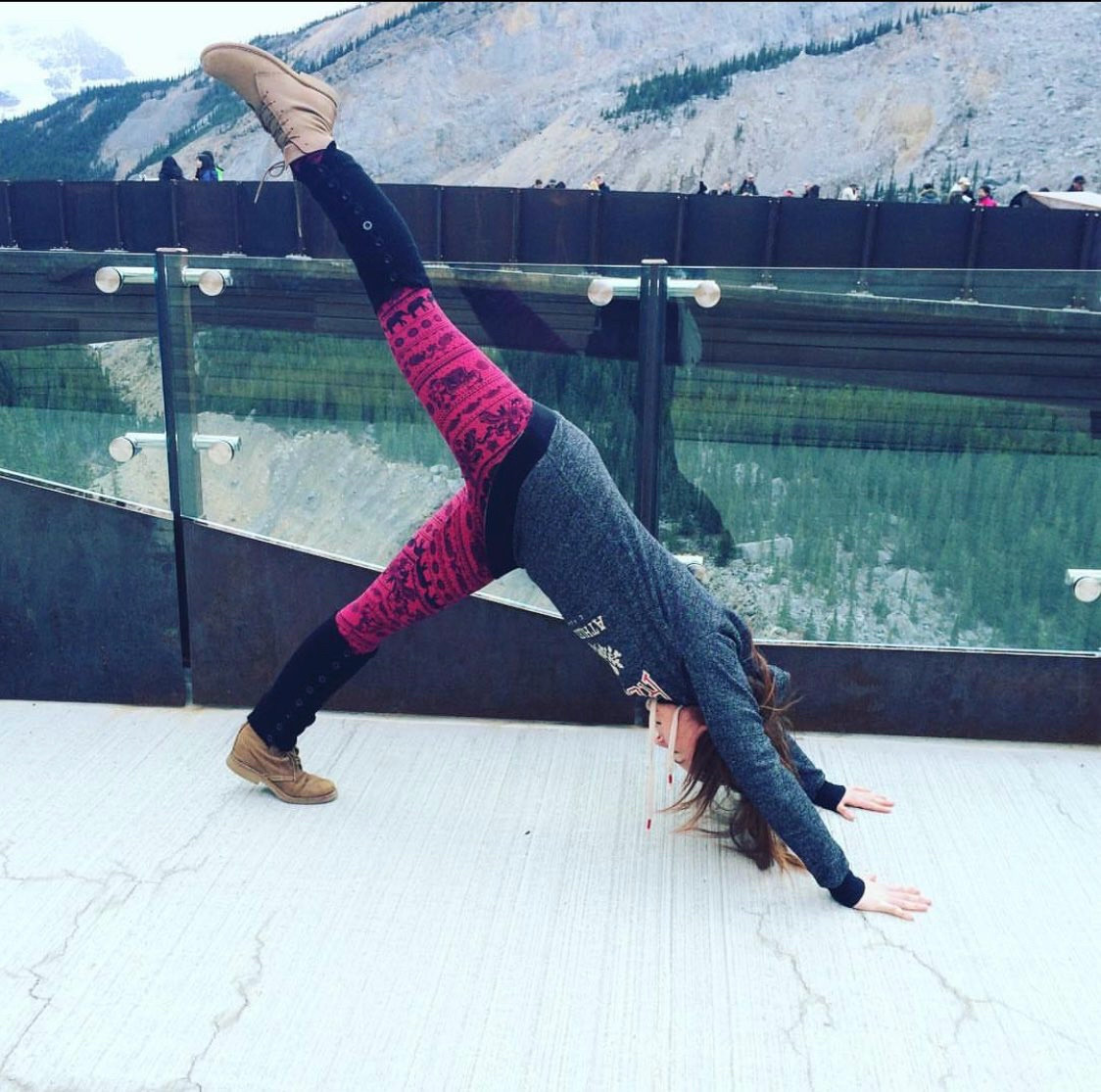 Kelly attempts the standing split yoga pose on the skywalk in Jasper National Park. She’s wearing red elephant leggings and black leg warmers and a grey sweater. There are mountains behind here and a glass wall showing a gorge below.