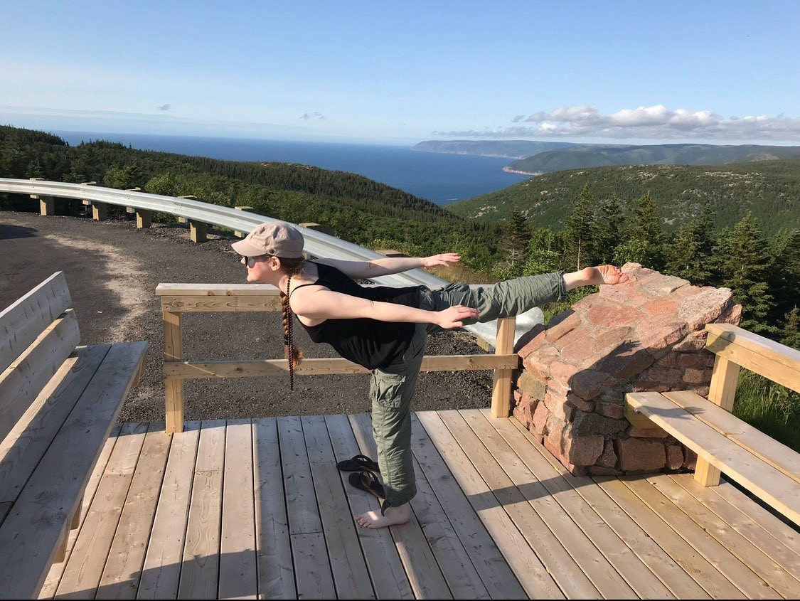 Kelly doing Warrior III yoga pose in Cape Breton on the Cabot Trail. There’s green rolling mountains and the Atlantic Ocean behind her and she’s barefoot wearing green pants, black tank and a grey hat.