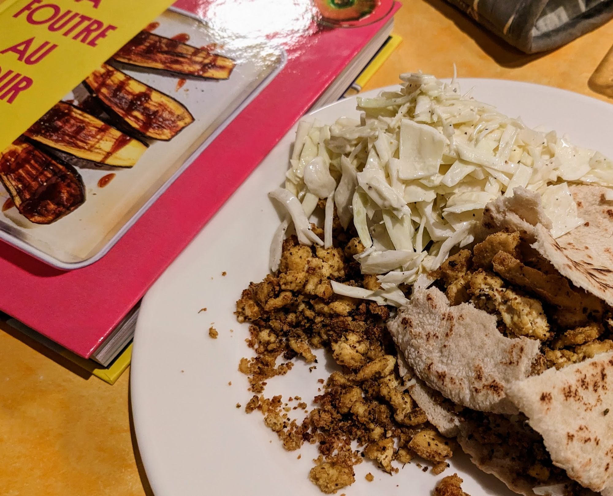 Une assiette avec des petits morceaux de tofu, du chou blanc cru et une pita dans un état assez déconstruit, avec un exemplaire du livre À Foutre Au Four derrière