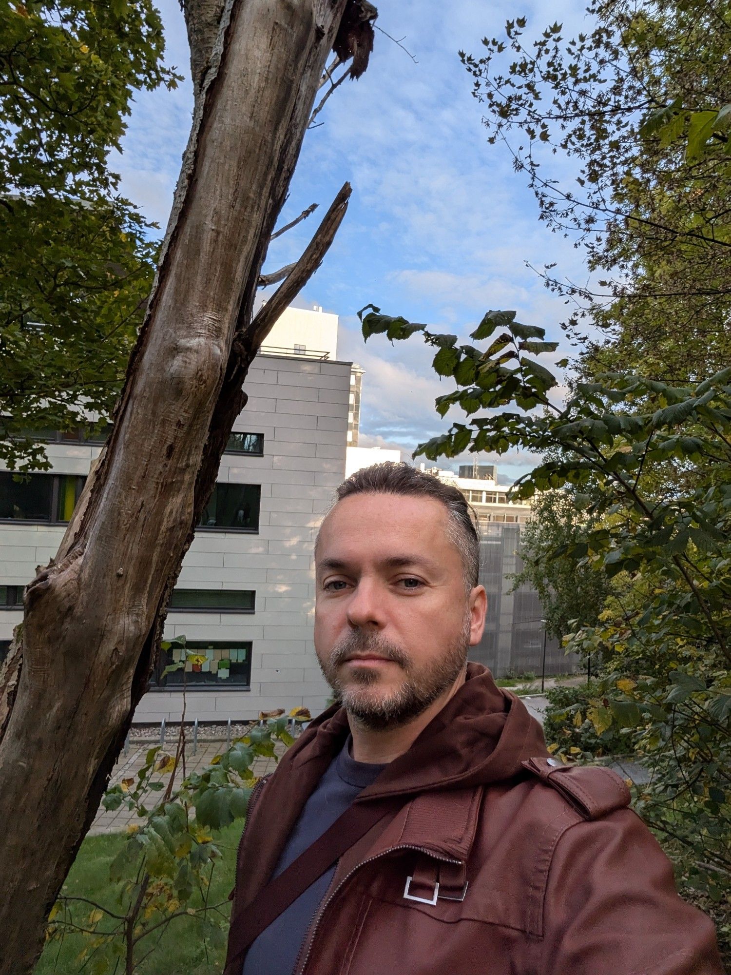 A man in early middle age (me) standing in a quiet part of the city with trees and a blue sky. I'm wearing a Burgundy jacket