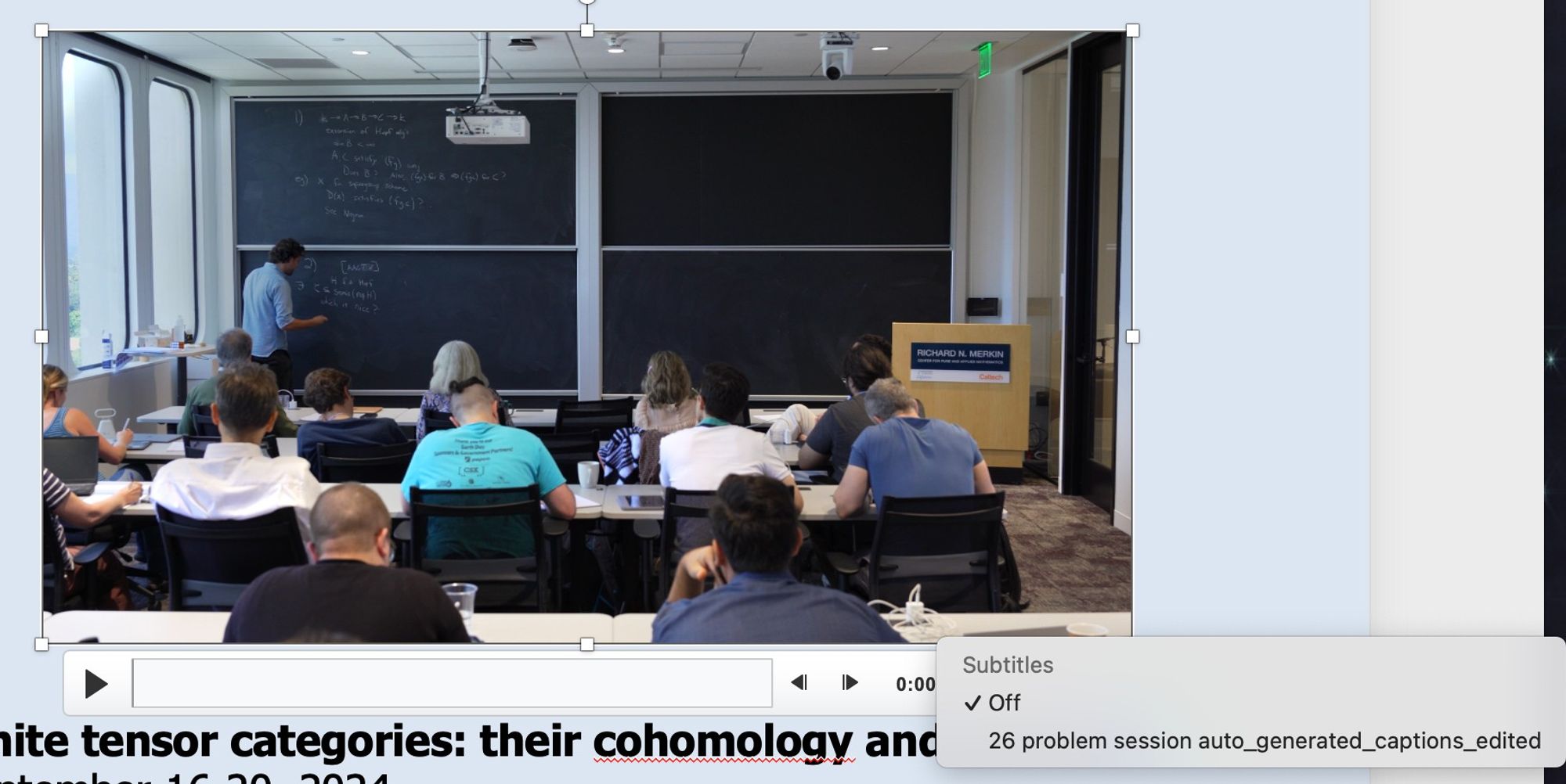 Screen shot of a powerpoint slide showing a video. There's a room of people facing a blackboard & one person writing on the board. Below the video is a bar with a play button on the left. On the right is a menu "Subtitles, Off, 26 problem session auto_generated_captions_edited"