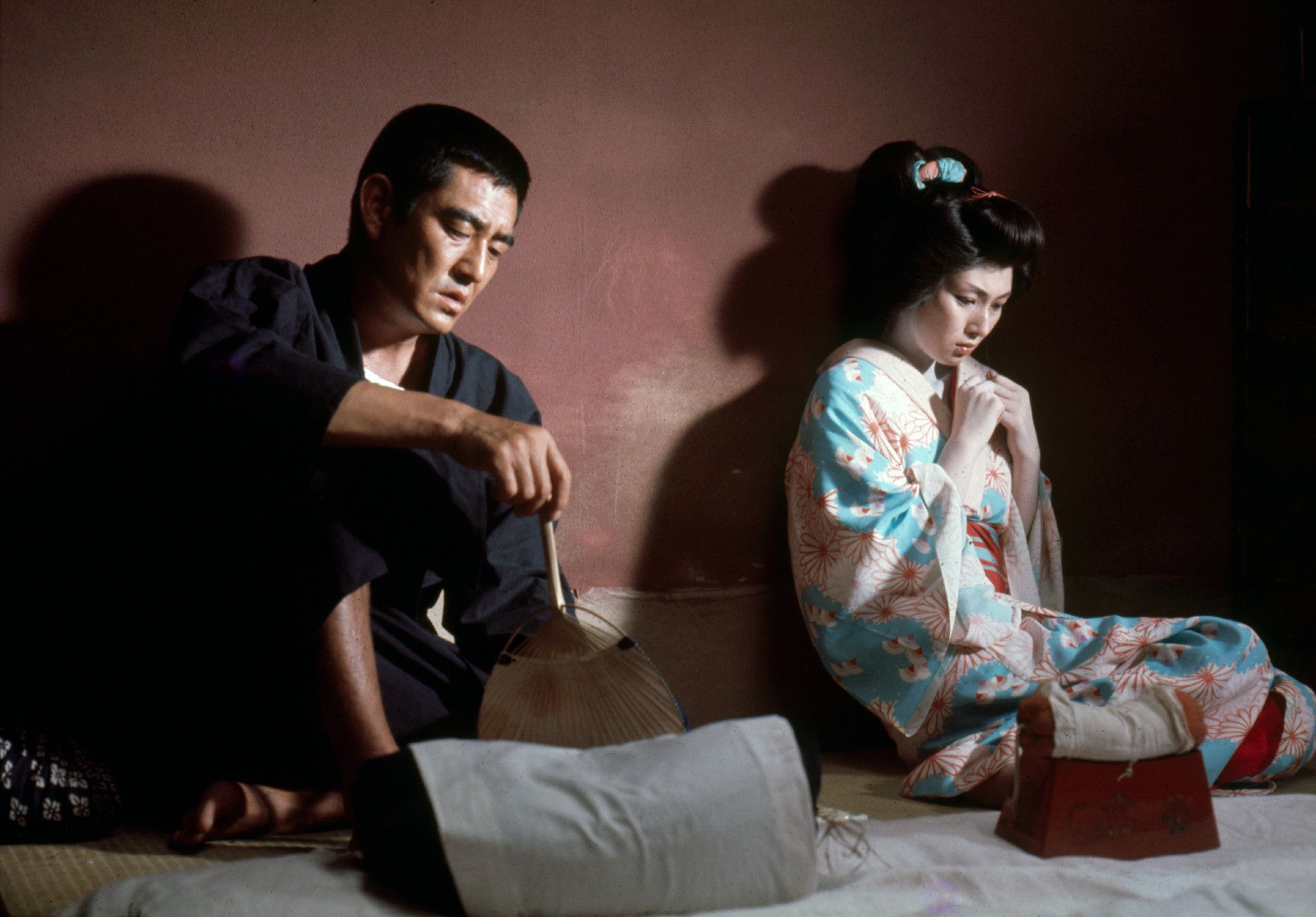 Meiko Kaji (梶芽衣子) and Ken Takakura (高倉 健) in The Homeless (無宿), 1974, directed by Koichi Saito (斉藤耕一). They're both sitting on the floor against a wall. Ken is holding a fan and looking down at it, while Meiko is wearing a kimono and kind of facing away from him, listening but guarded/uncomfortable.