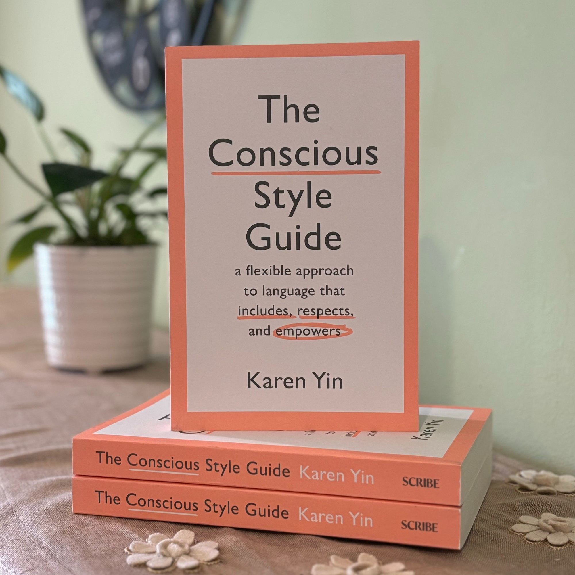 Photo of The Conscious Style Guide standing on two other copies, stacked. The cover is off-white, with a coral border. The words conscious, includes, and respects are underlined; the word empowers is circled. In the background, a small leafy plant in a white pot beside a large wall clock.