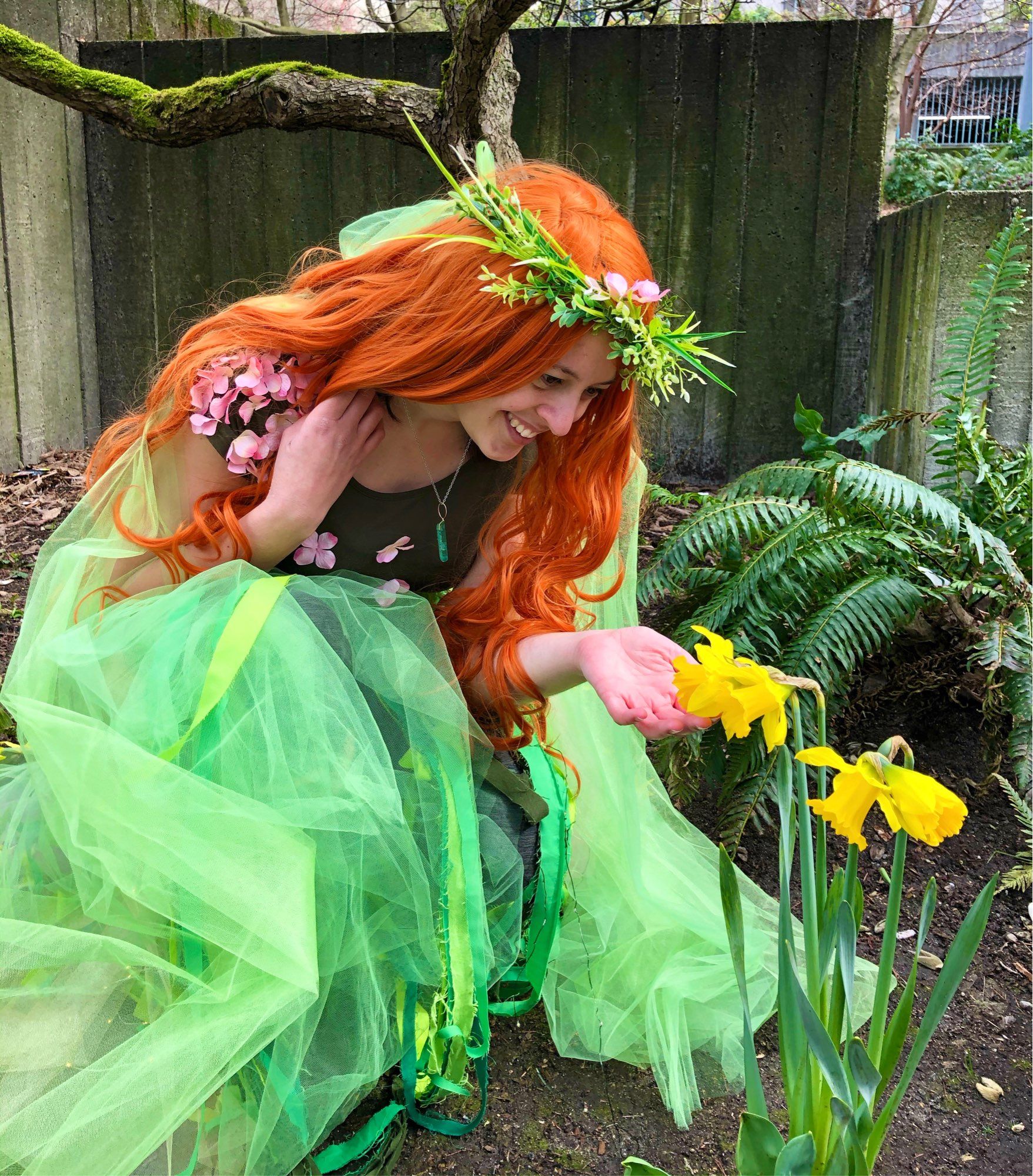 Cosplay of a nature spirit, long red-orange hair, lots of green bits of fabric, kneeling down next to some daffodils and smiling at them