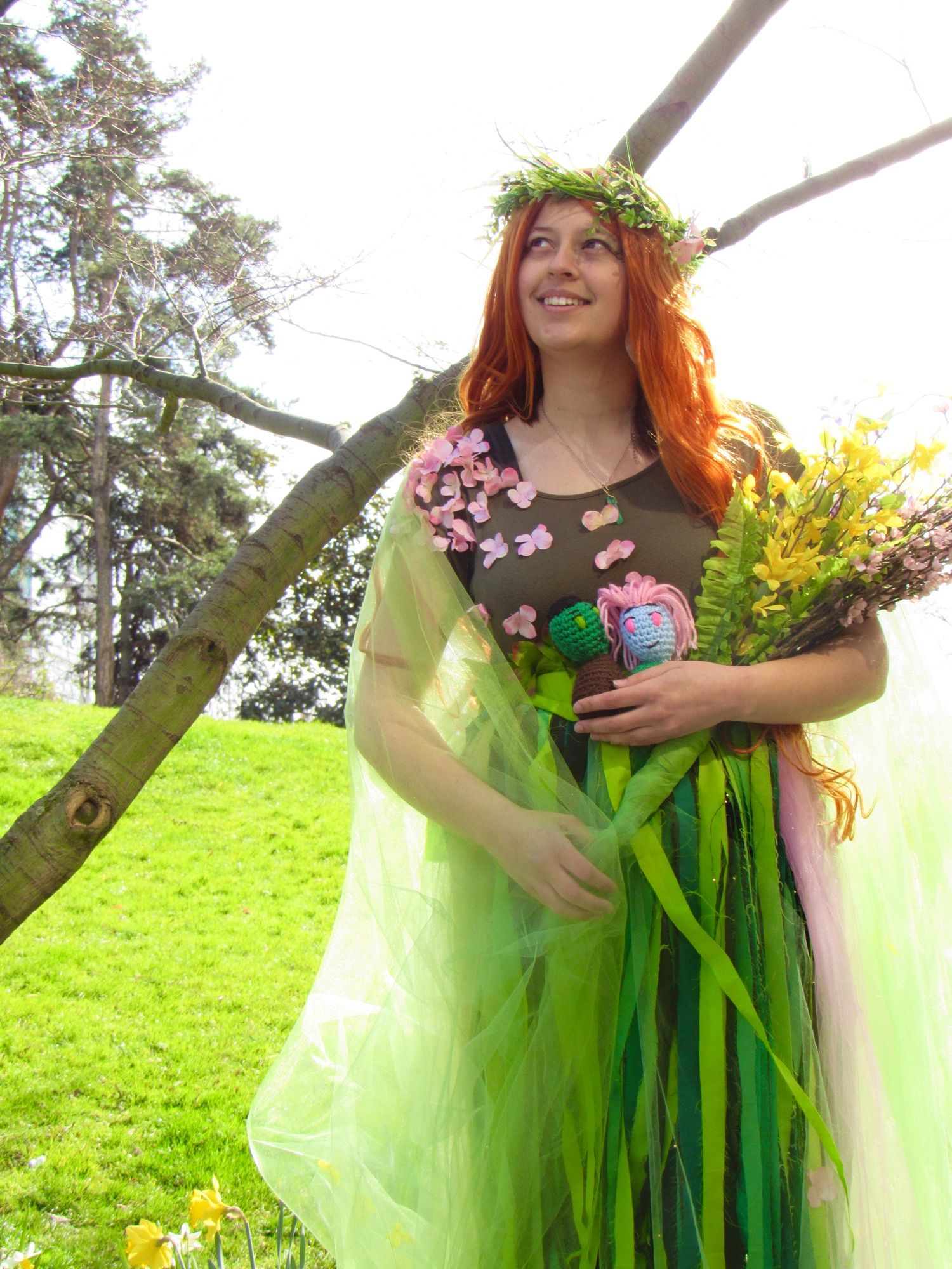 Cosplay of the Wildmother from Critical Role. Lots of green fabric, dress, and carrying tiny plushies of Fjord and Caduceus. Looking into the distance smiling 