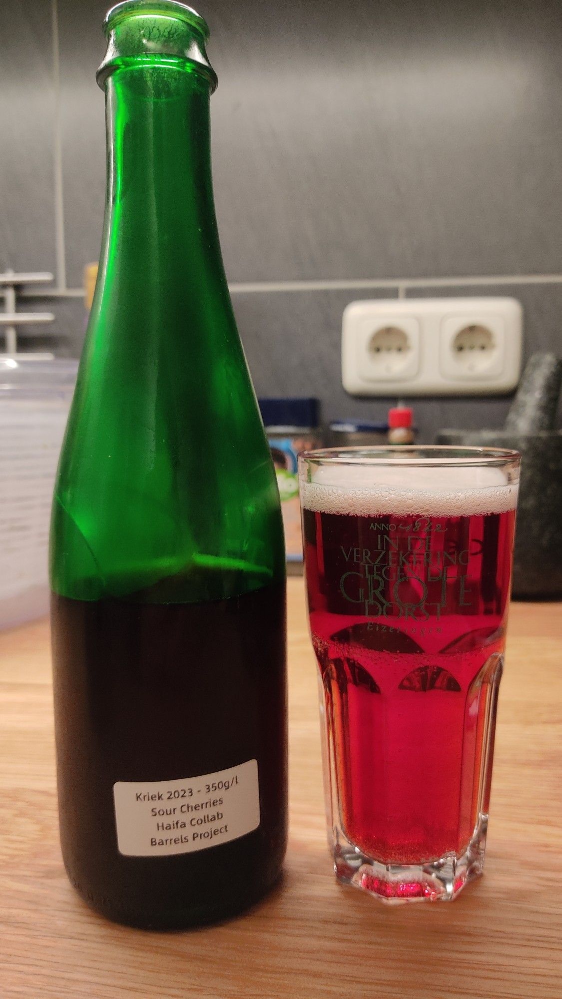 A glass with red beer, next to a lambic bottle labeled "Haifa Barrel Collab Sour Cherry Kriek 2023"