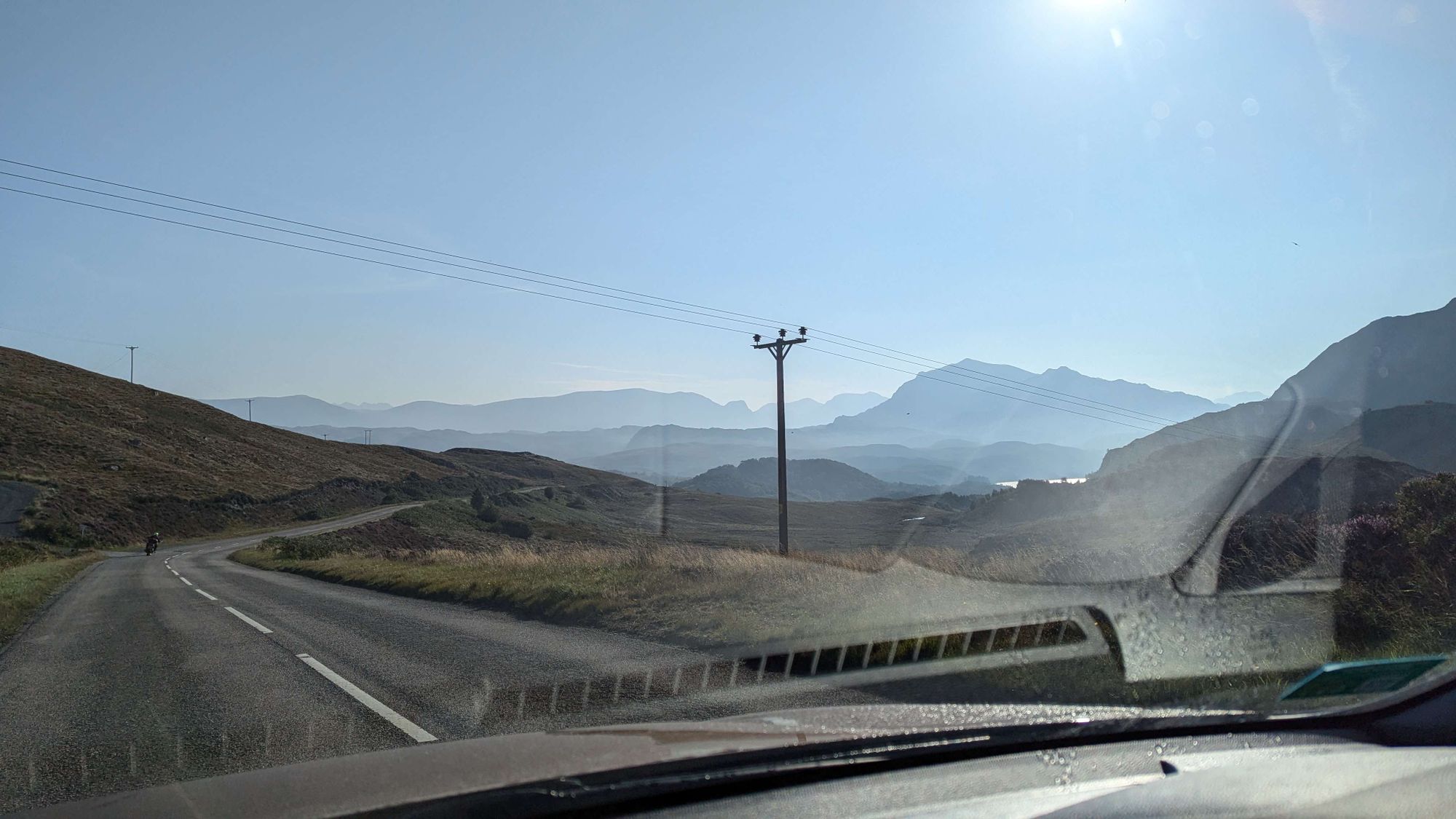 Windschutzscheibenperspektive: Blauer Himmel, weite Landschaft mit Bergketten im Hintergrund, die durch Dunst unterschiedlich verdeckt werden. Im Vordergrund kreuzt eine Stromleitung die Straße.