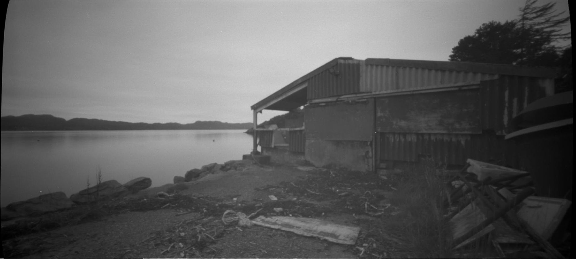 Schwarzweißaufnahme, düster. Blick auf einen desolaten Schuppen rechts, links Wasser.