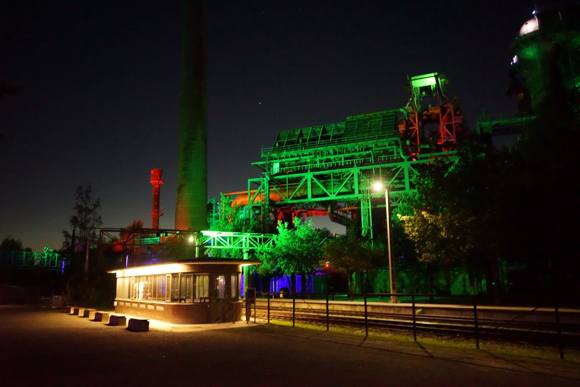 Nachtaufnahme, links ein flaches normal gelblich beleuchtetes Gebäude, rechts im Hintergrund grün blau und rot beleuchtete Industrieanlagen.