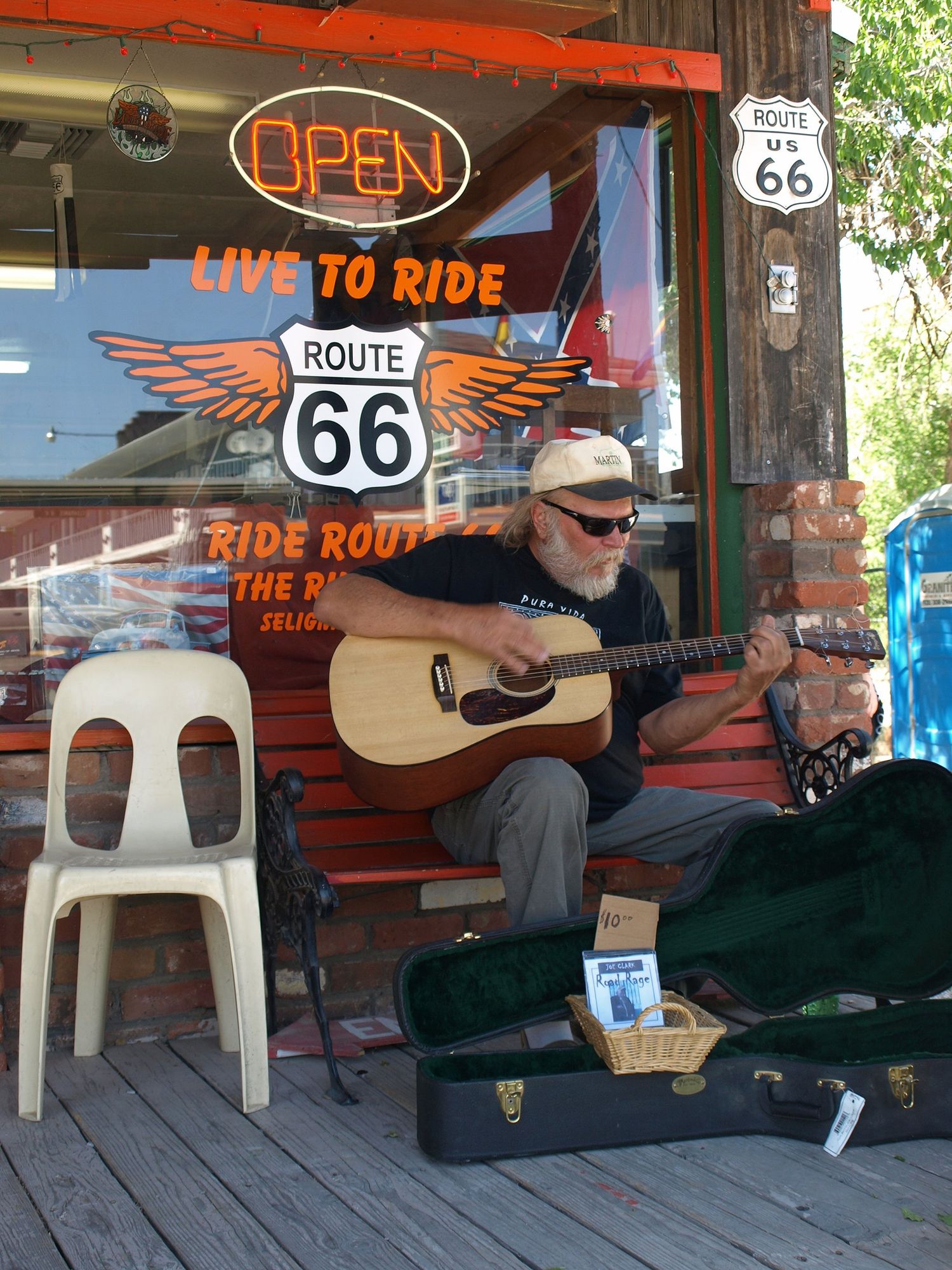 Vor einem Cafe mit orangener Fensterbeschriftung "Route 66 Live to ride" sitzt auf einer Holzbank ein Gitarrist mit akustischer Gitarre und schwarzem T-Shirt.