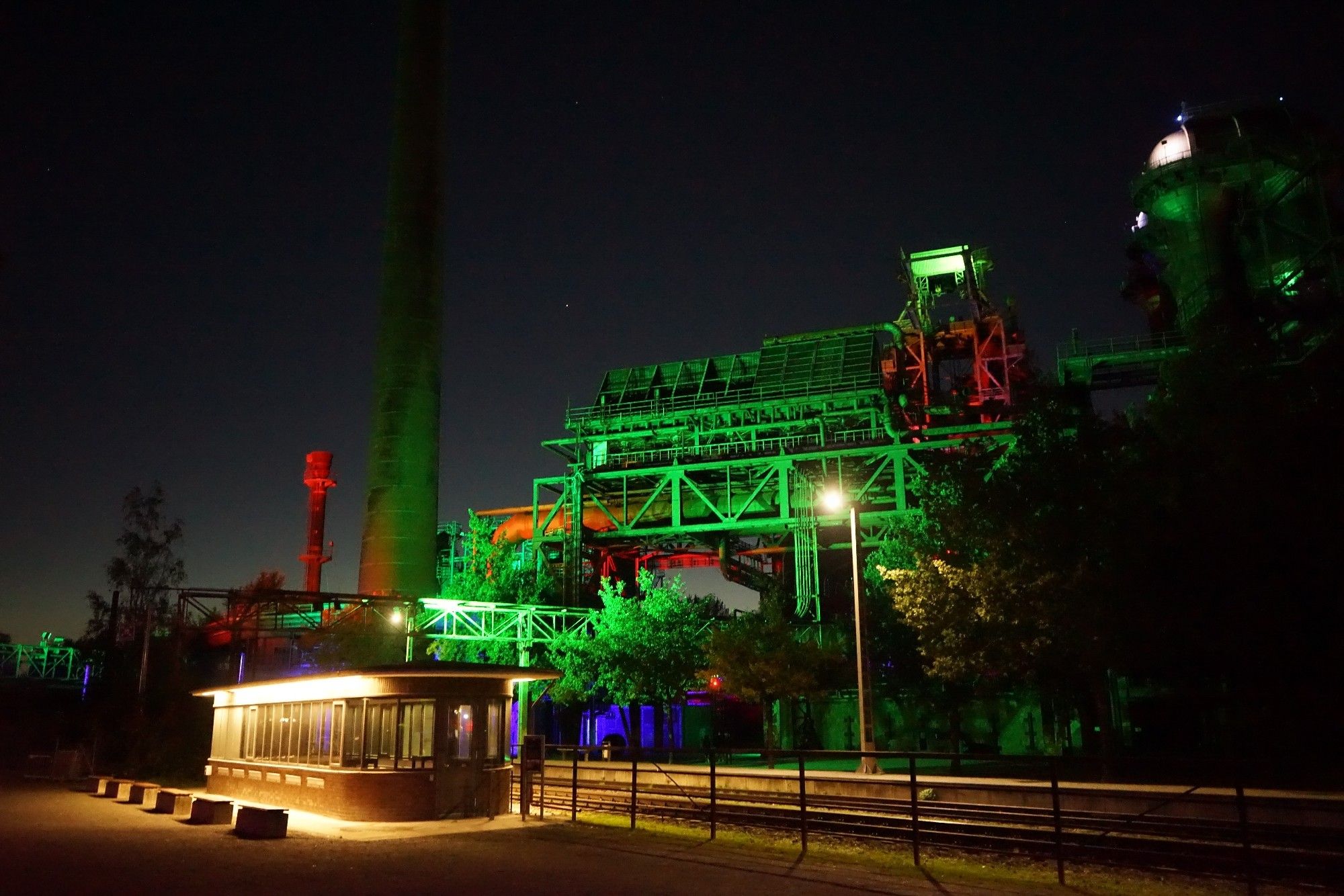 Nachtaufnahme, links ein flaches normal gelblich beleuchtetes Gebäude, rechts im Hintergrund grün blau und rot beleuchtete Industrieanlagen.