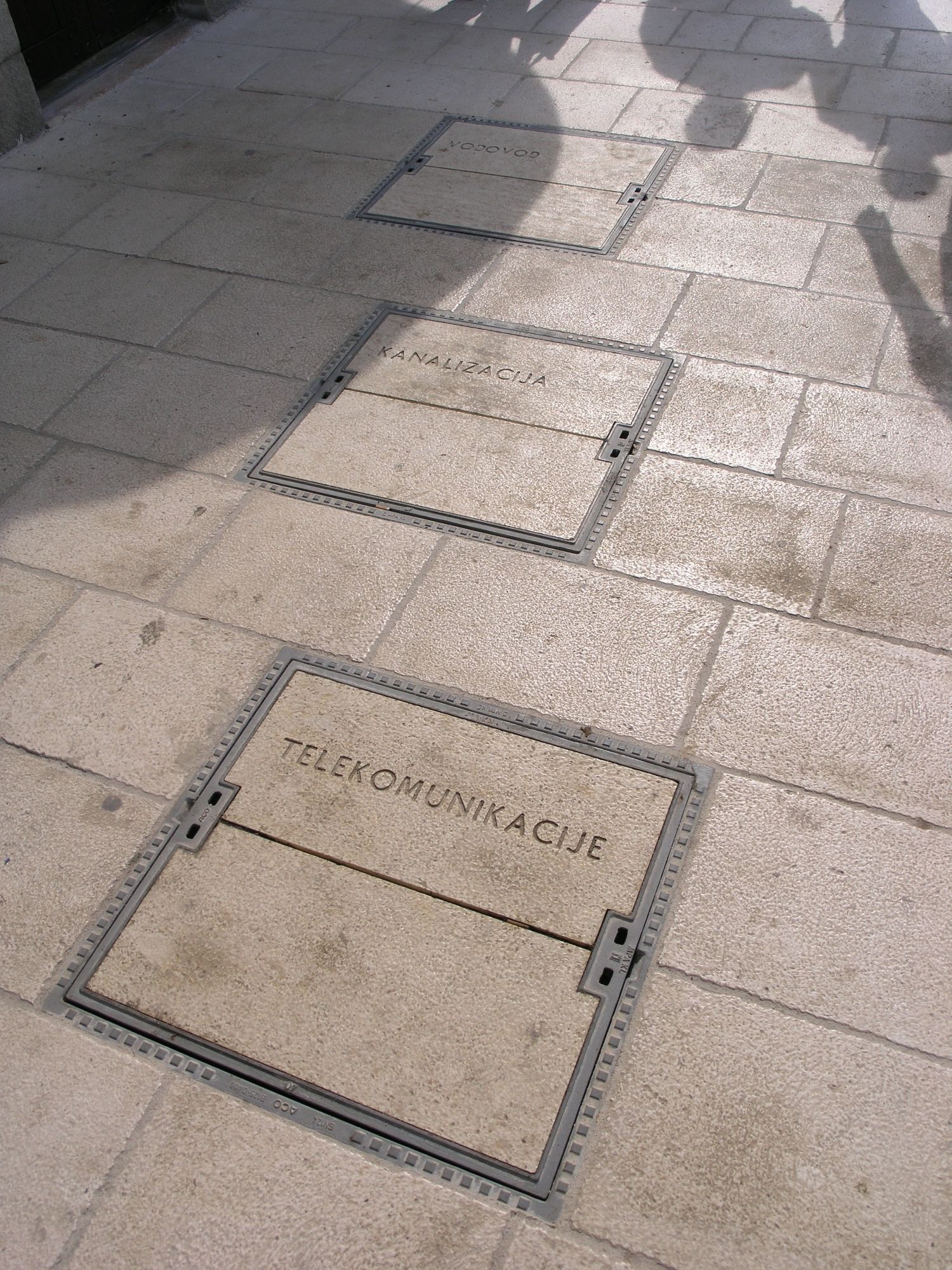 Quadratischer Schachtdeckel, schräg aufgenommen. In den Beton eingelassen der Schriftzug "TELEKOMUNIKACIJE". Dahinter, leicht versetzt zwei weitere Schachtdeckel gleicher Bauweise mit Beschriftung "KANALIZACIJE und VODOVOD"