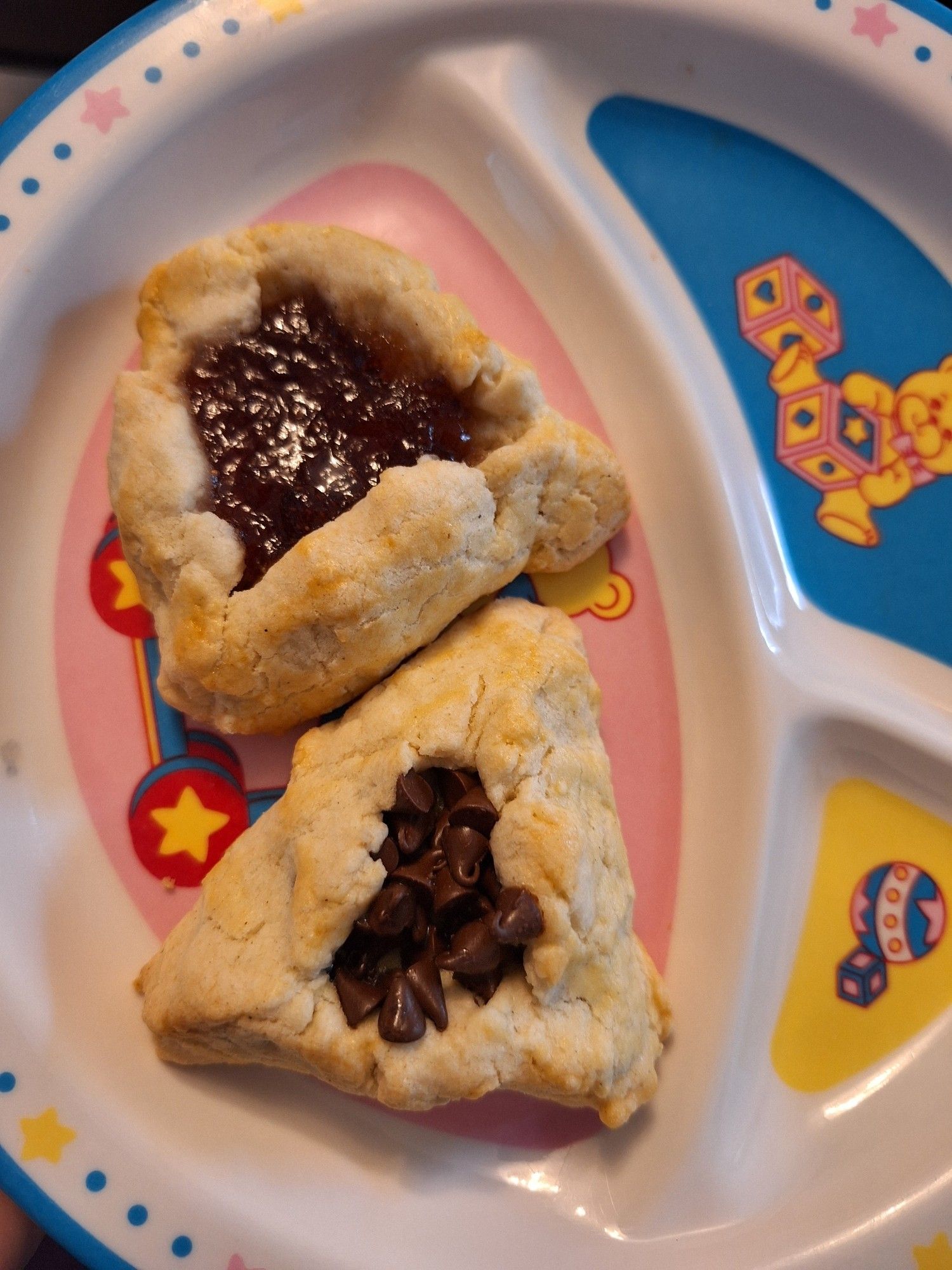 Two hamantaschen - triangular shortbread cookies with filling - on a child's plate. One is filled with strawberry jelly, the other with chocolate chips.