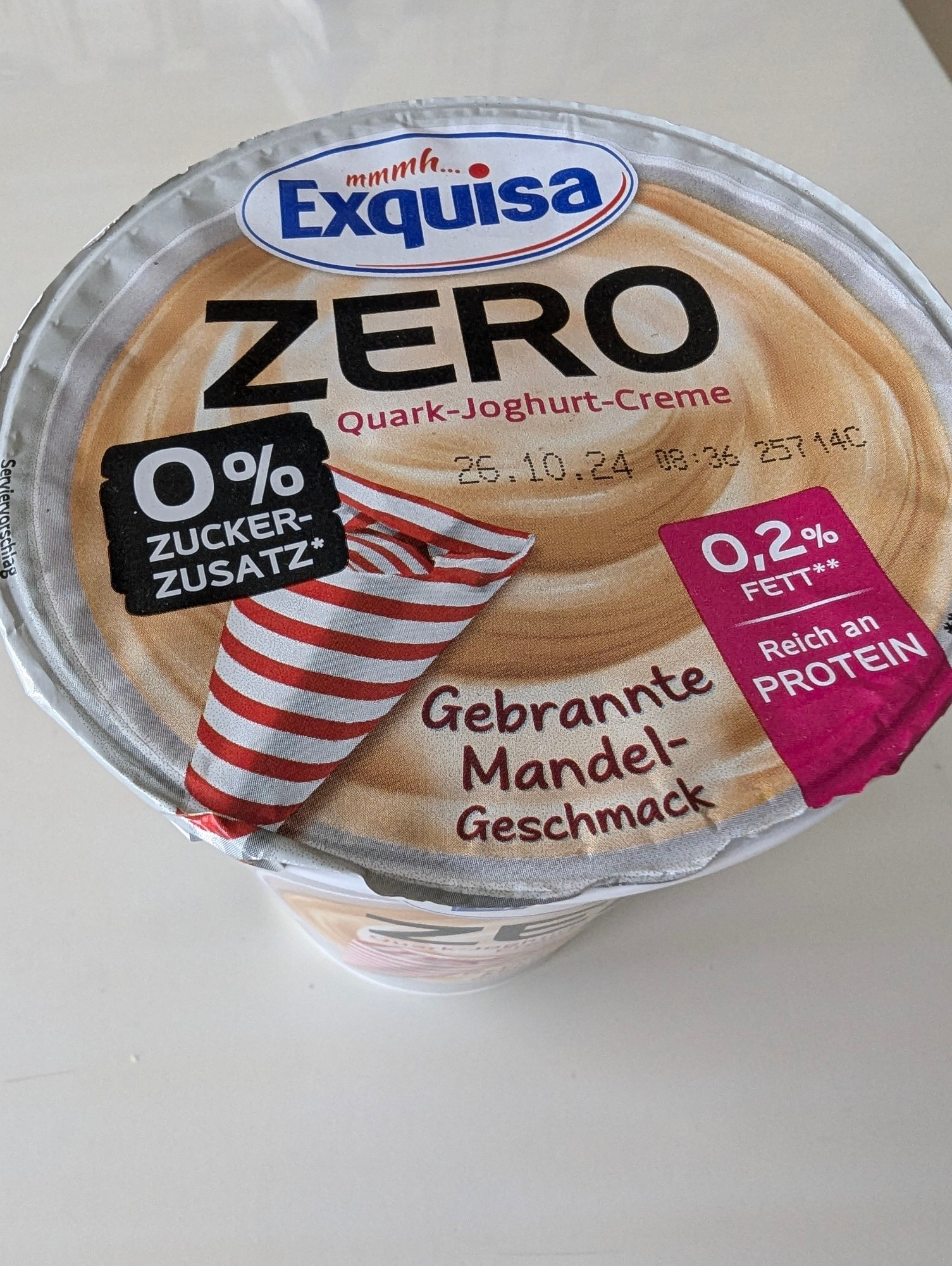 Das Bild zeigt einen Behälter mit Exquisa “ZERO” Quark mit Joghurtcreme. Auf der Verpackung steht, dass das Produkt keinen Zuckerzusatz und nur 0,2% Fett enthält. Es ist reich an Proteinen und hat den Geschmack “Gebrannte Mandel”. Das Mindesthaltbarkeitsdatum ist der 25. Oktober 2024.