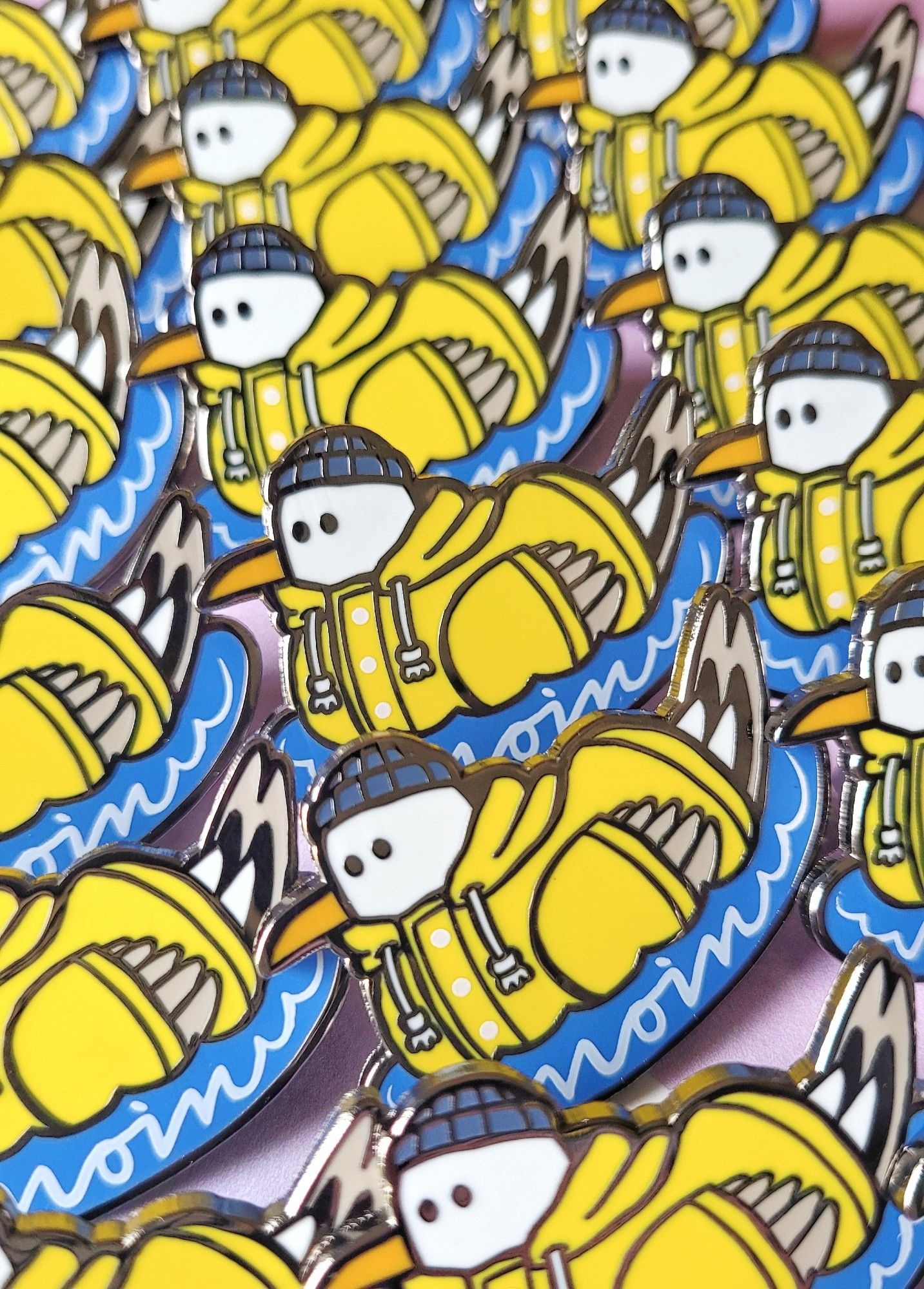 Photo of enamel pins of a seagull wearing a yellow raincoat and a blue hat, floating on blue water. The word "moin", a common greeting in north germany, is screen printed on the water.